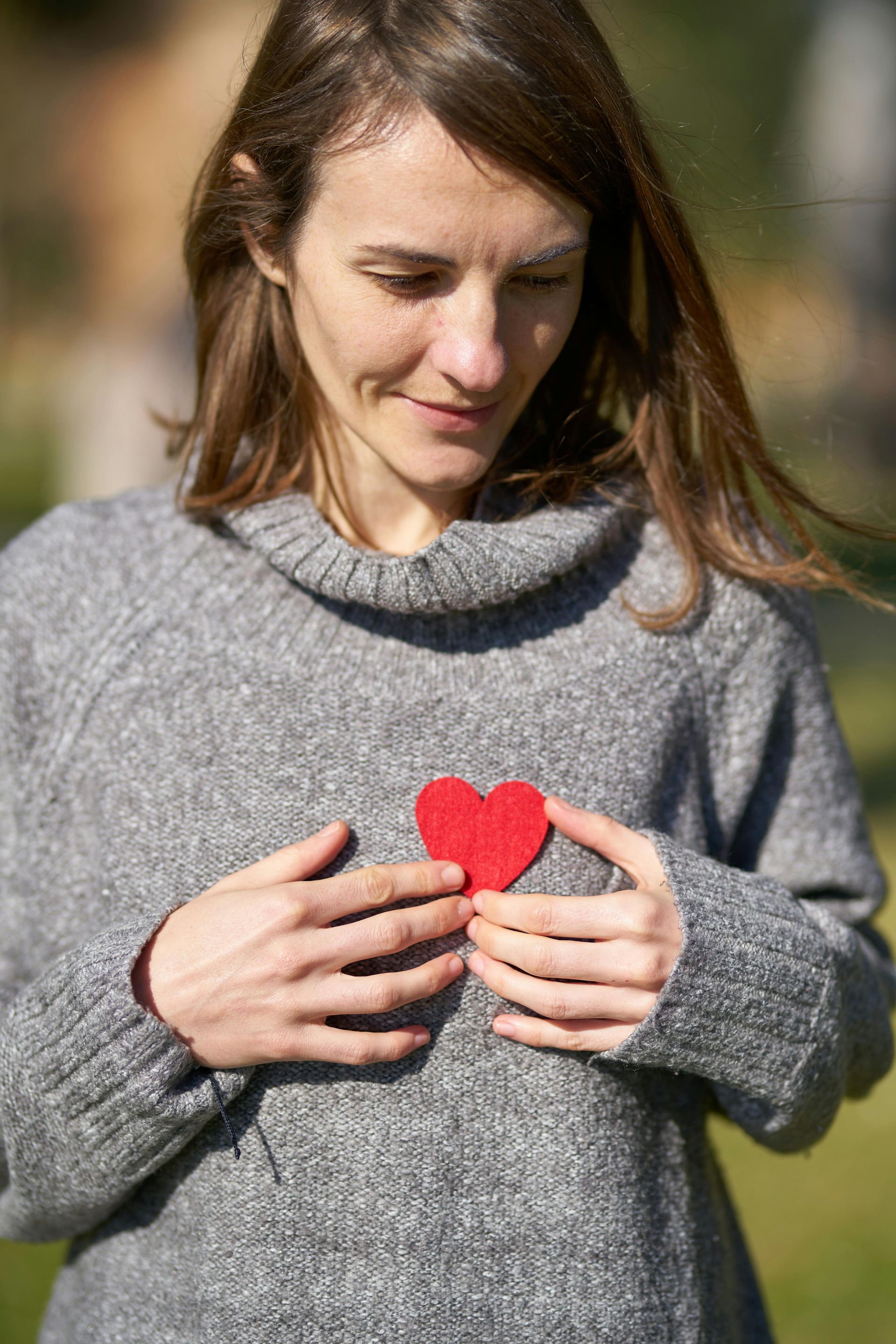 Woman holding a red heart over her chest; wearing a gray sweater, smiling outdoors.