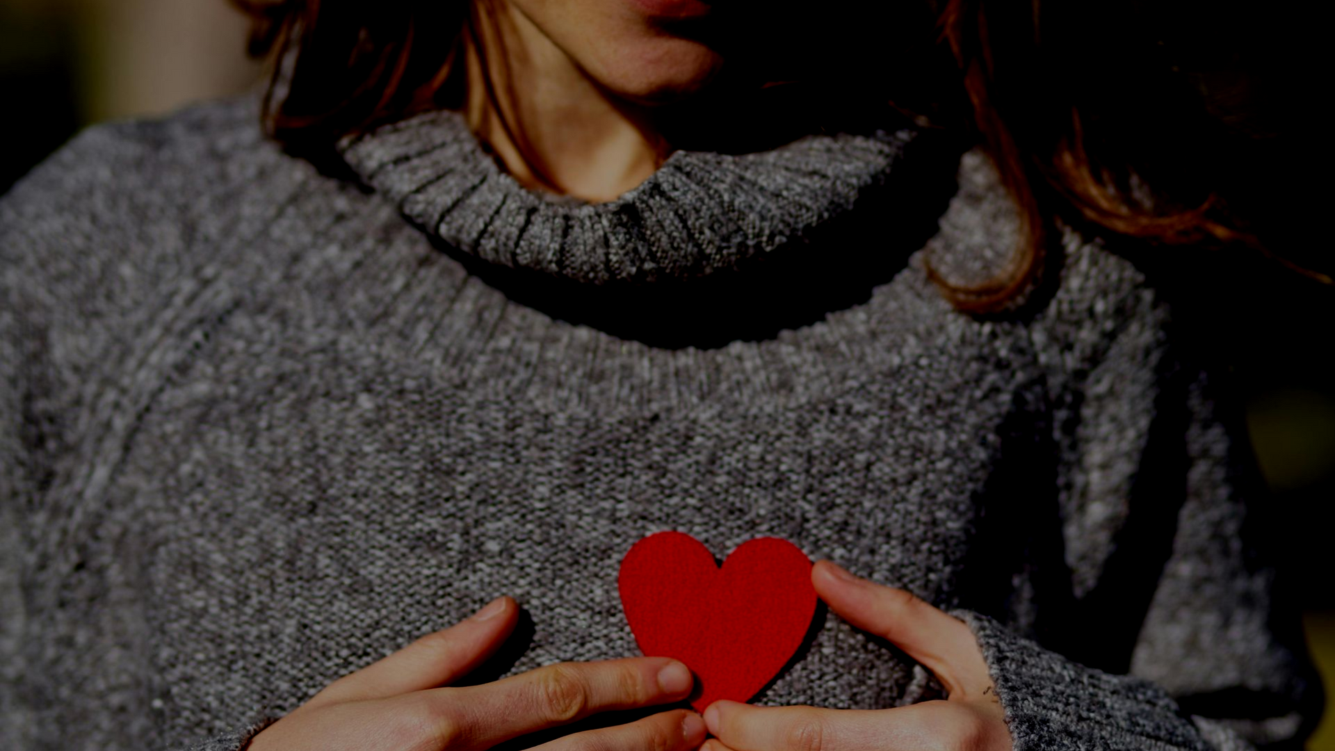 Woman in gray sweater holding a red paper heart over her chest.