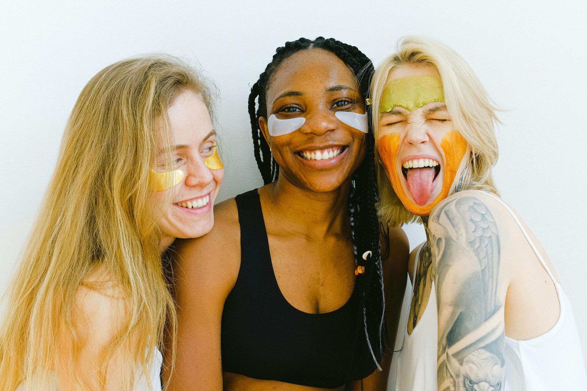 Three diverse women smiling, wearing face masks. Blonde woman sticks out tongue, other two laugh. White background.