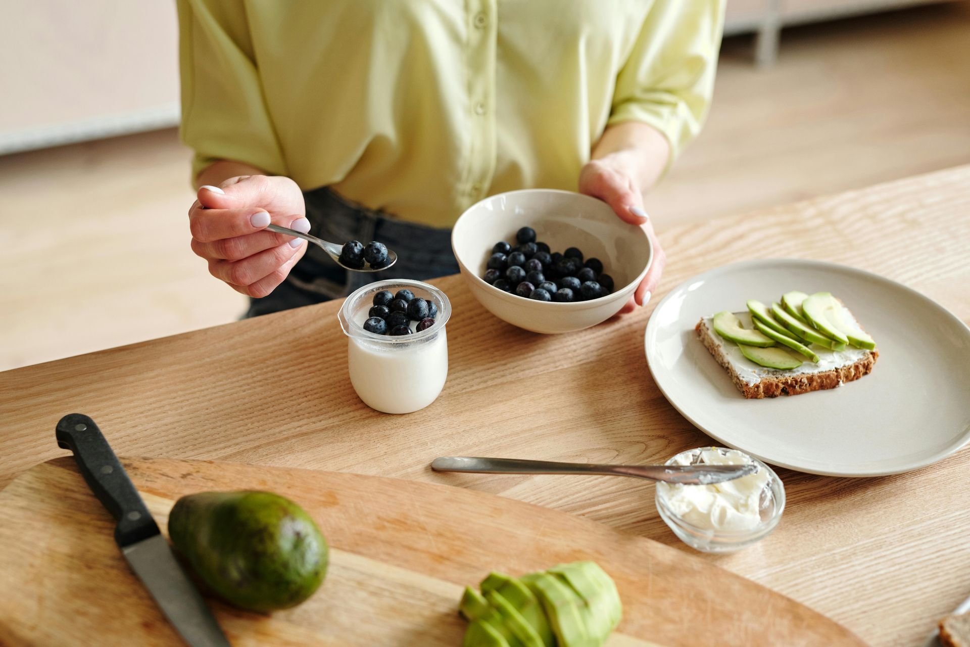 Woman prepares a healthy snack: yogurt with blueberries, avocado toast. Wooden table setting.