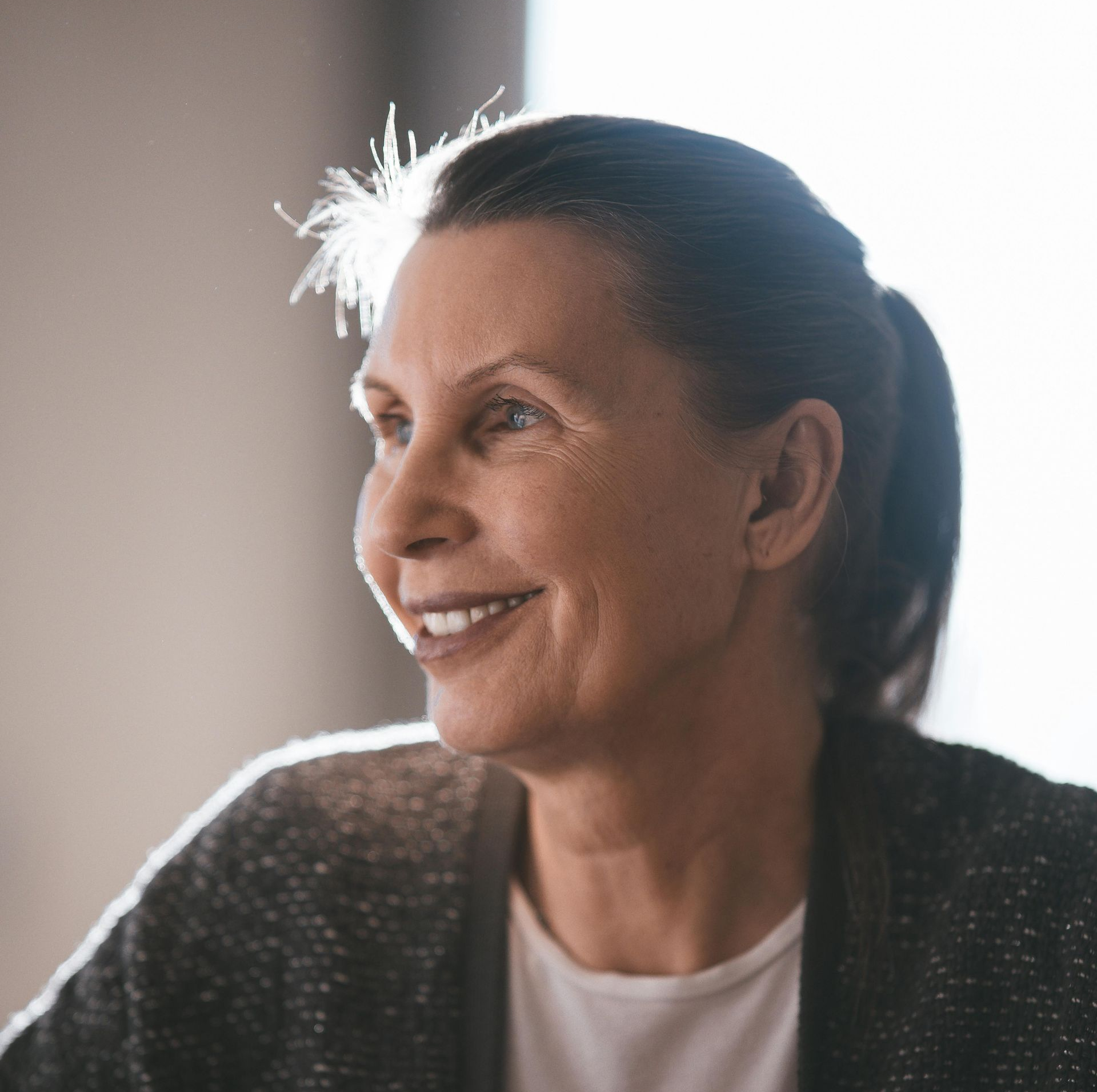Woman with dark hair in a ponytail, smiling, wearing a dark cardigan, with bright background.