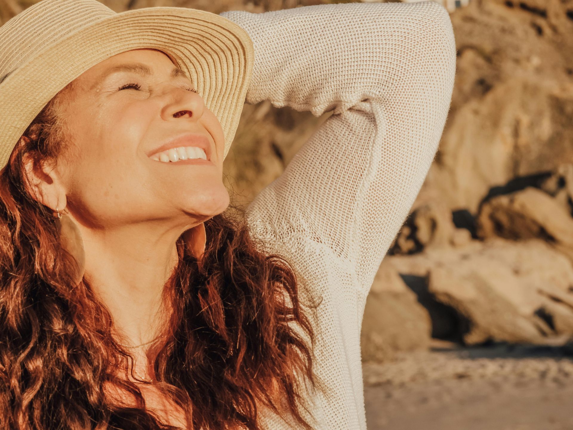 Woman in a sunhat smiles, looking upwards with arm raised, on a beach.