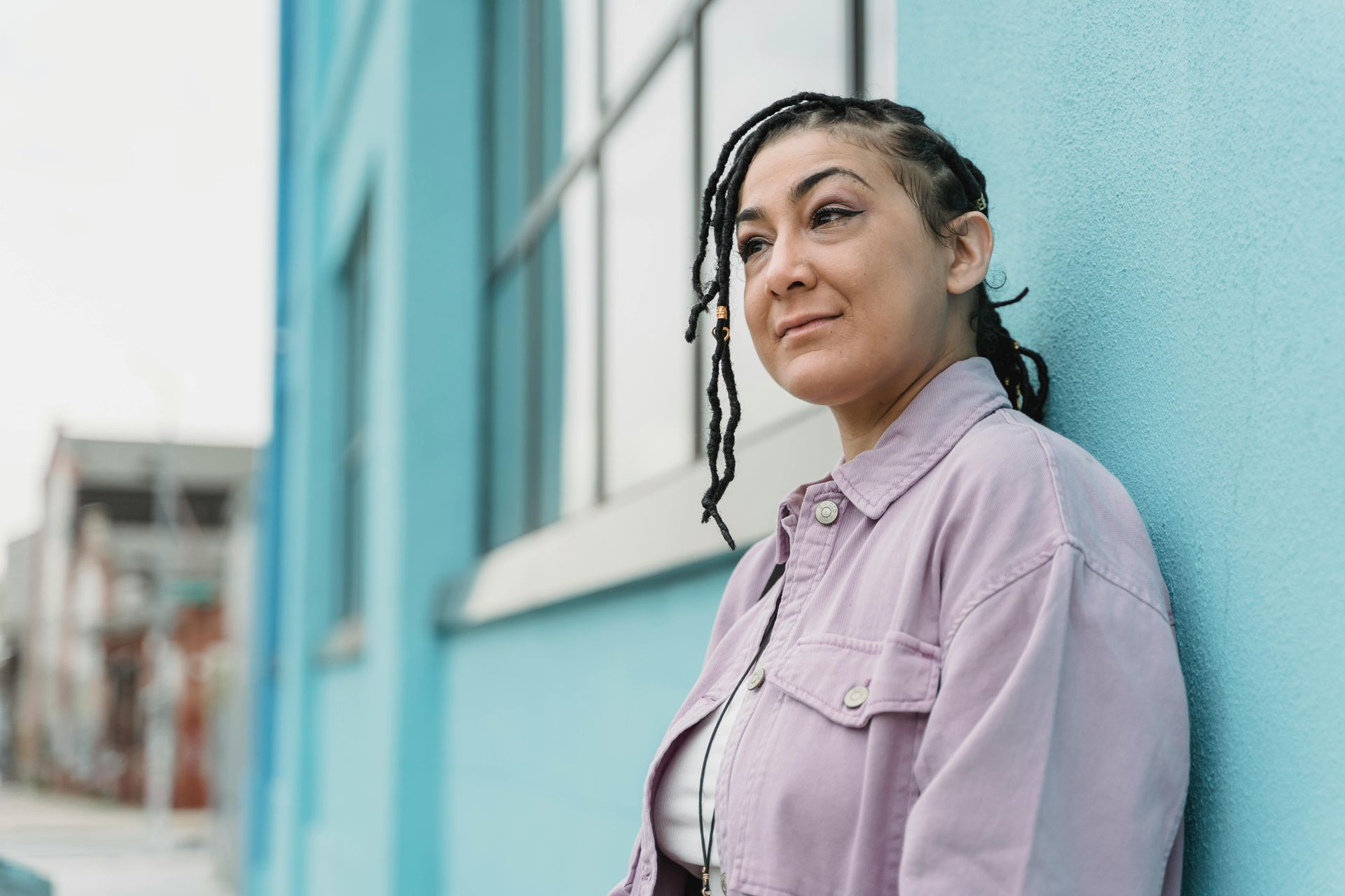 Woman with braided hair leans against a light blue building, wearing a lavender jacket, looking thoughtful.