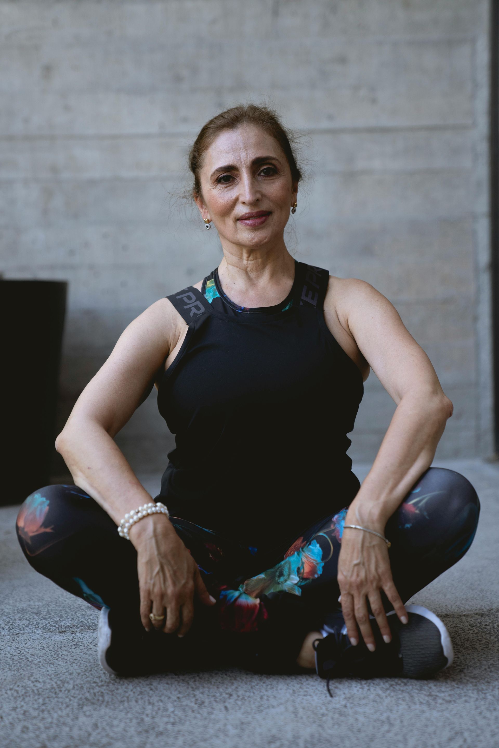 Woman in workout clothes seated cross-legged, smiling, outdoors.