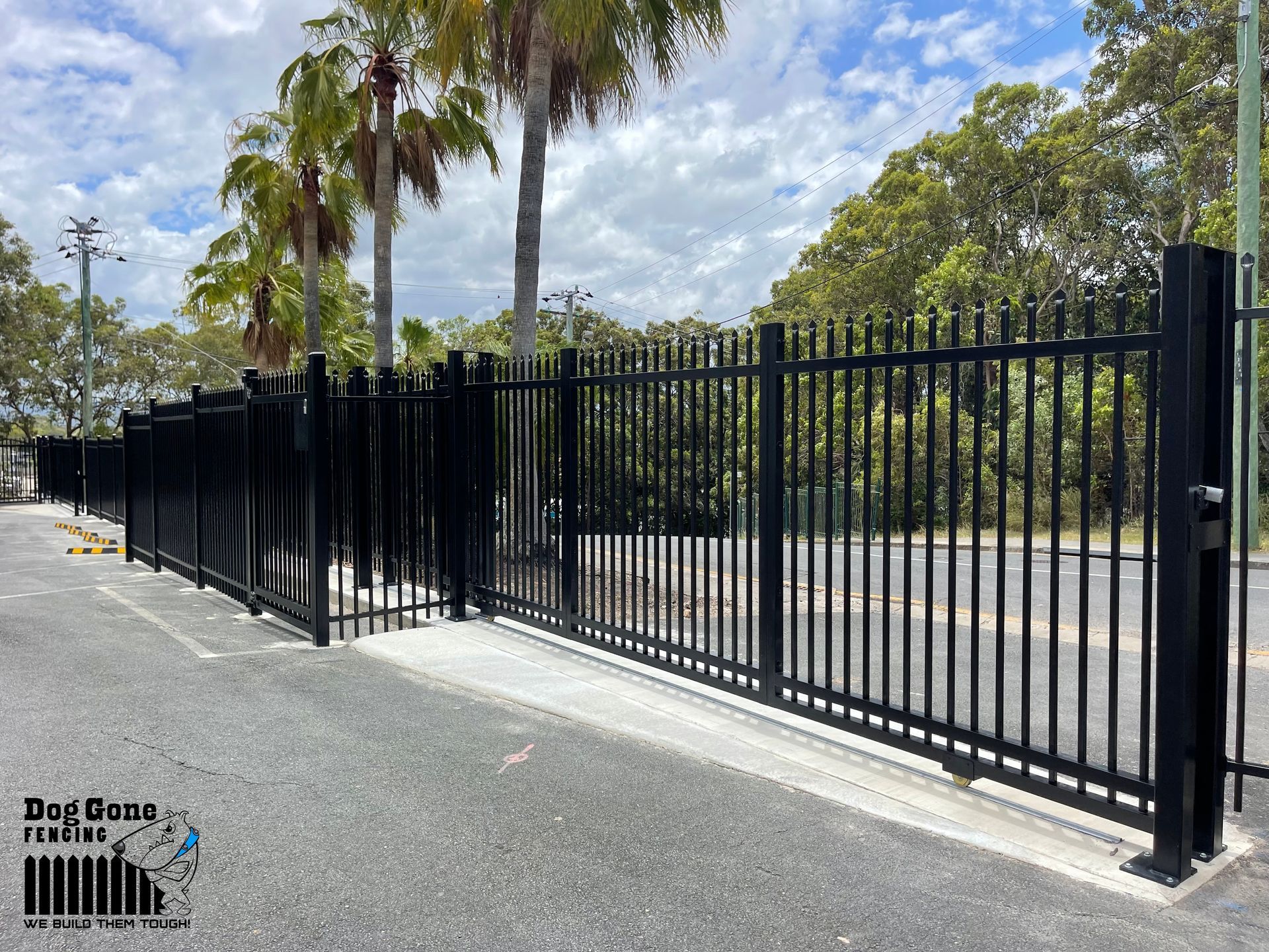 A Black Metal Fence Surrounds A Parking Lot With Palm Trees In The Background — Dog Gone Fencing in Paget, QLD