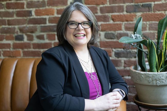 Woman with glasses, smiling, in a black blazer, sitting by a brick wall and a potted plant.
