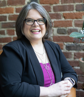 A woman wearing glasses and a black jacket is smiling while sitting in front of a brick wall.