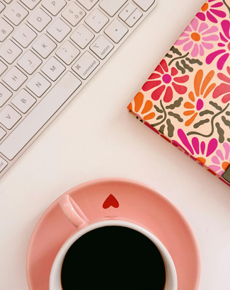 A cup of coffee on a saucer next to a keyboard and a floral-patterned notebook.