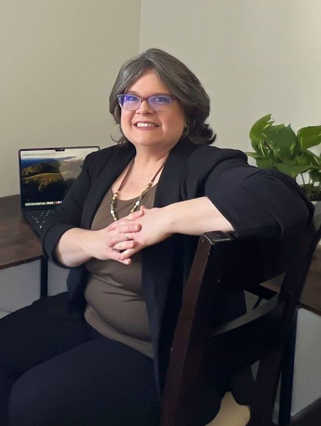 Woman with glasses, typing on a laptop at a desk, smiling. She wears a black blazer in a neutral-toned room.