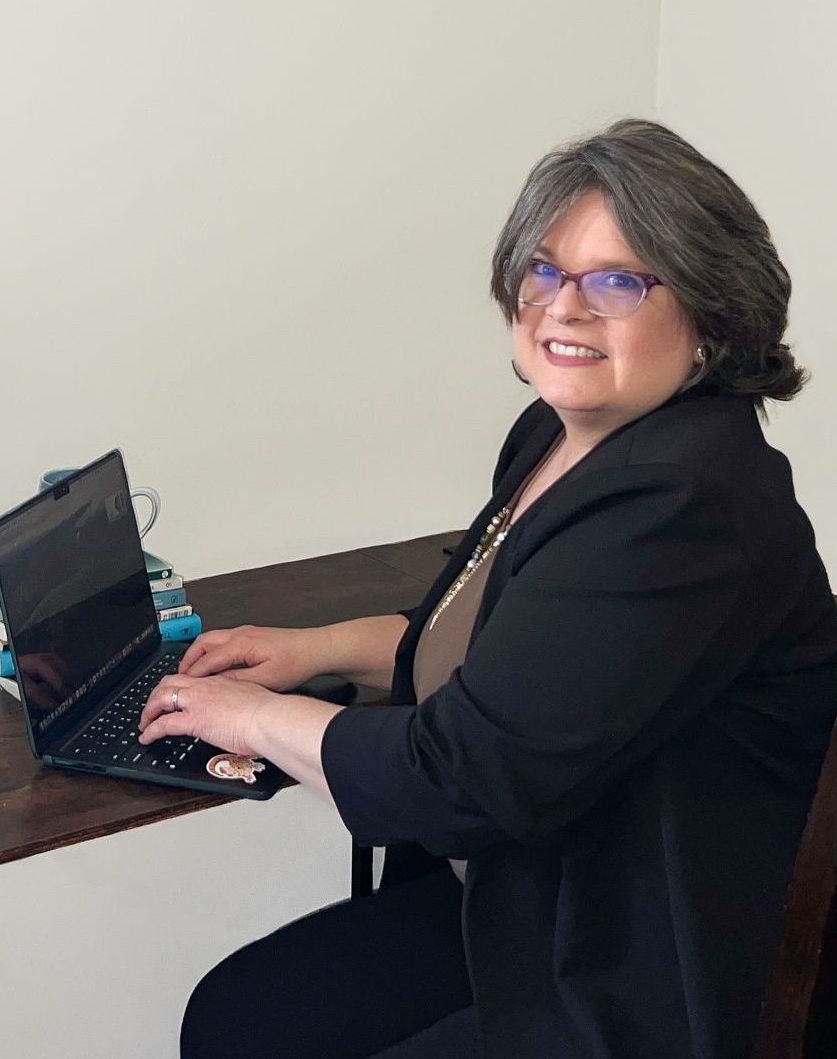 Woman with glasses, typing on a laptop at a desk, smiling. She wears a black blazer in a neutral-toned room.