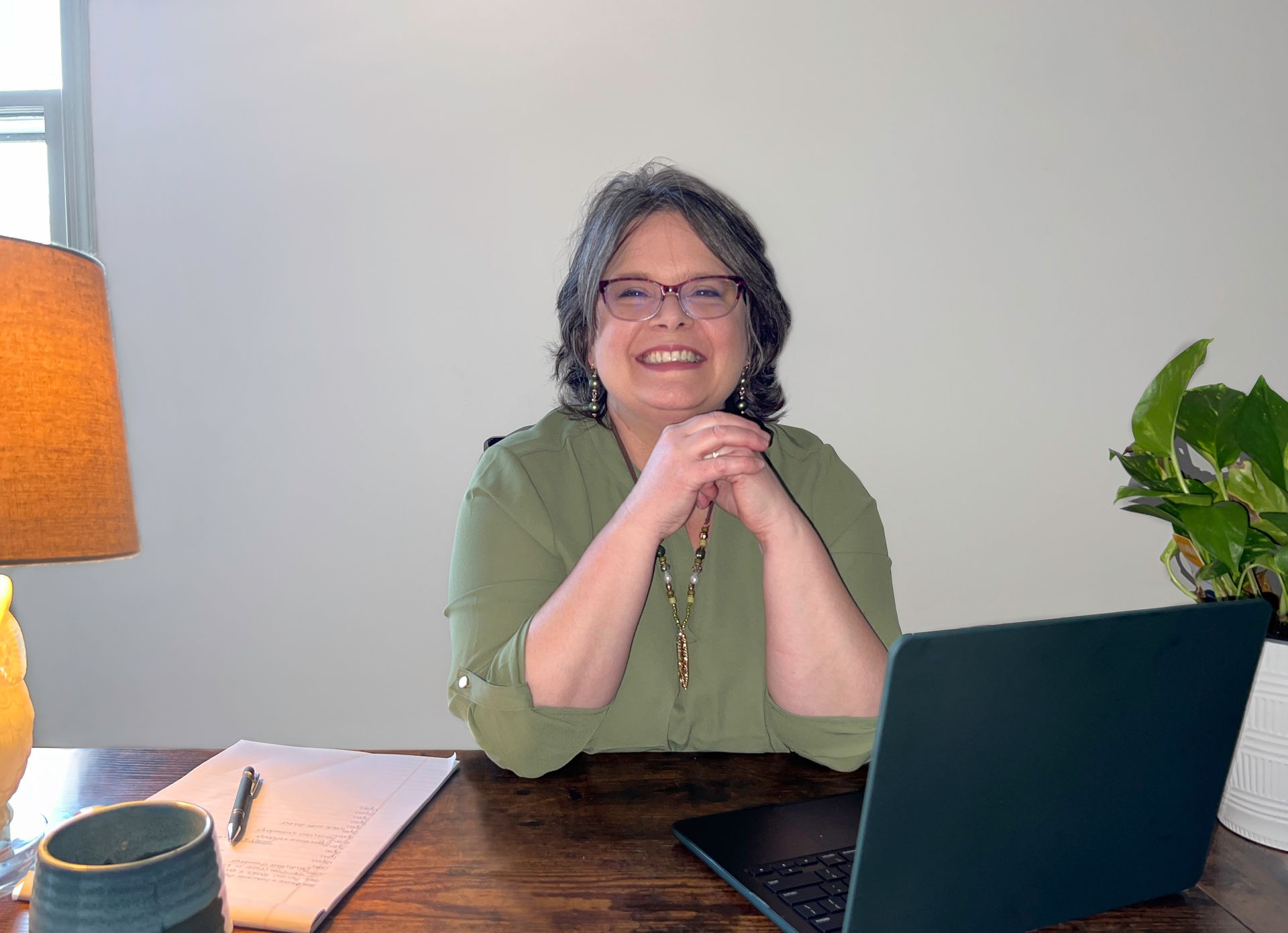 Woman with glasses smiles at the camera, seated at a desk with a laptop, papers, and a lamp. She is wearing a green shirt.