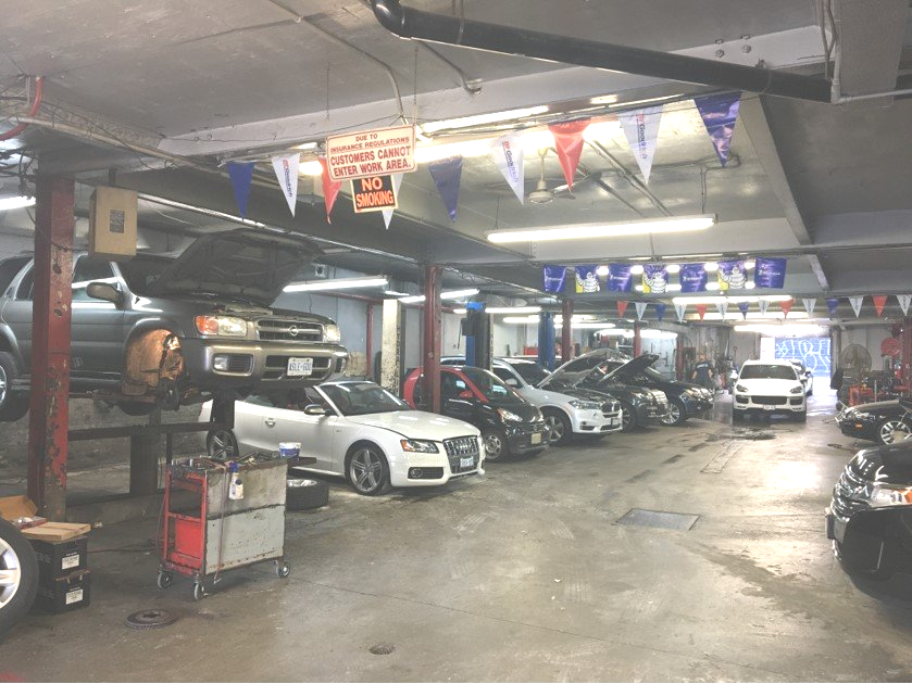 Cars in a garage, some on lifts. Mechanics working. Banners and tools visible.