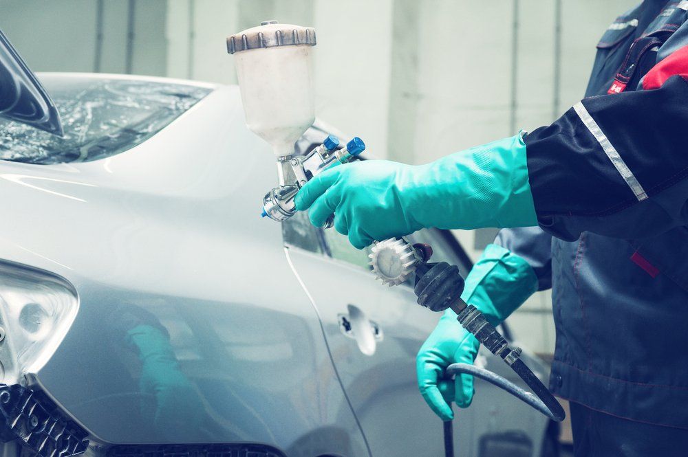 Person in gloves painting a car silver in a workshop with a spray gun.