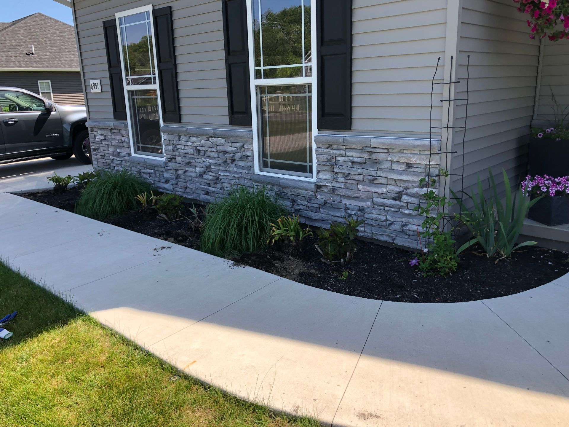 A gray house with black shutters features a stone-veneer foundation and a manicured flower bed along a concrete walkway.