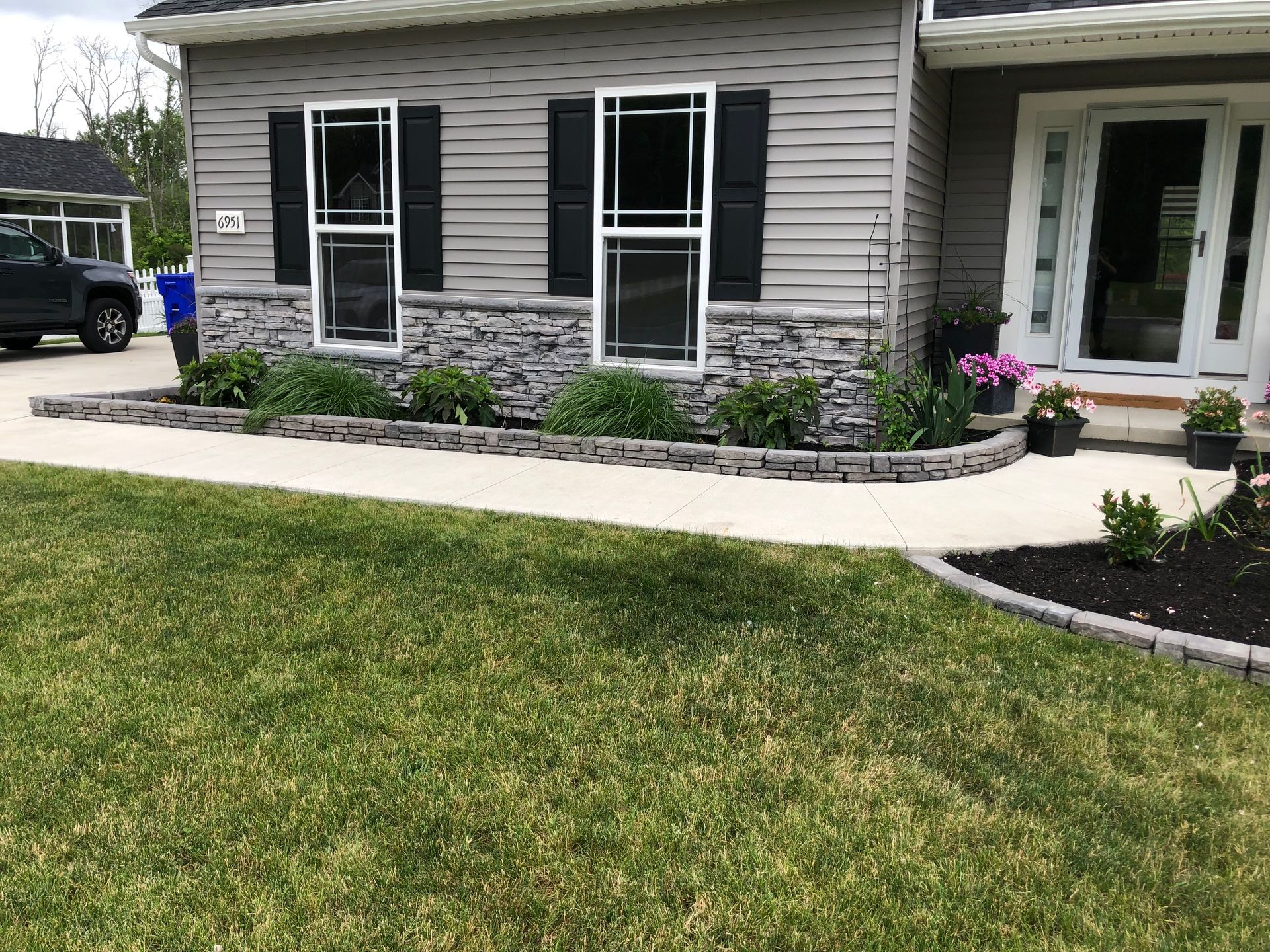 A concrete walkway curves along the front of a house with light gray siding, stone veneer, and green landscaping.