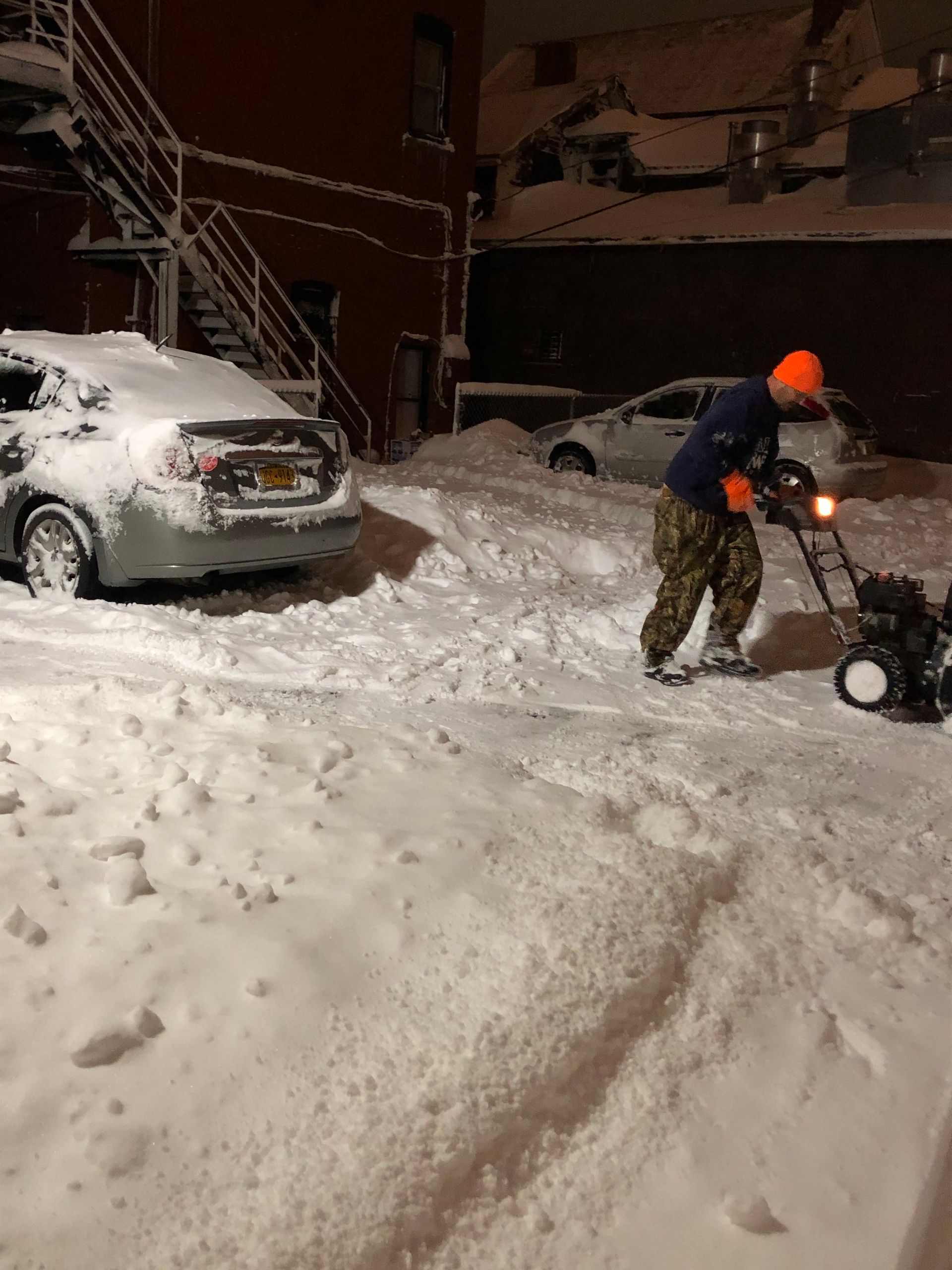 A person in an orange beanie and camouflage pants uses a snowblower to clear a snow-covered parking area at night.
