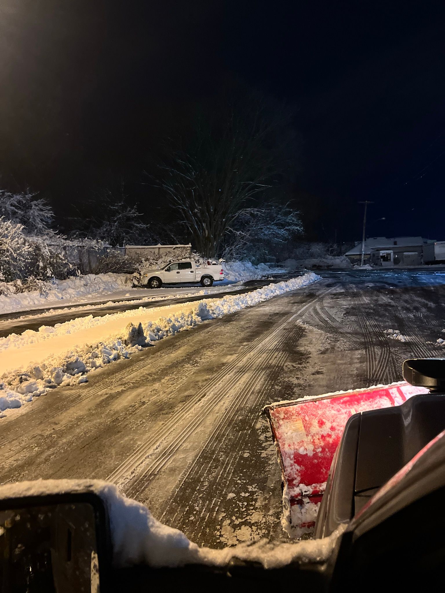 A view from inside a vehicle at night, showing a red snow plow blade clearing a snowy lot near a parked white truck.