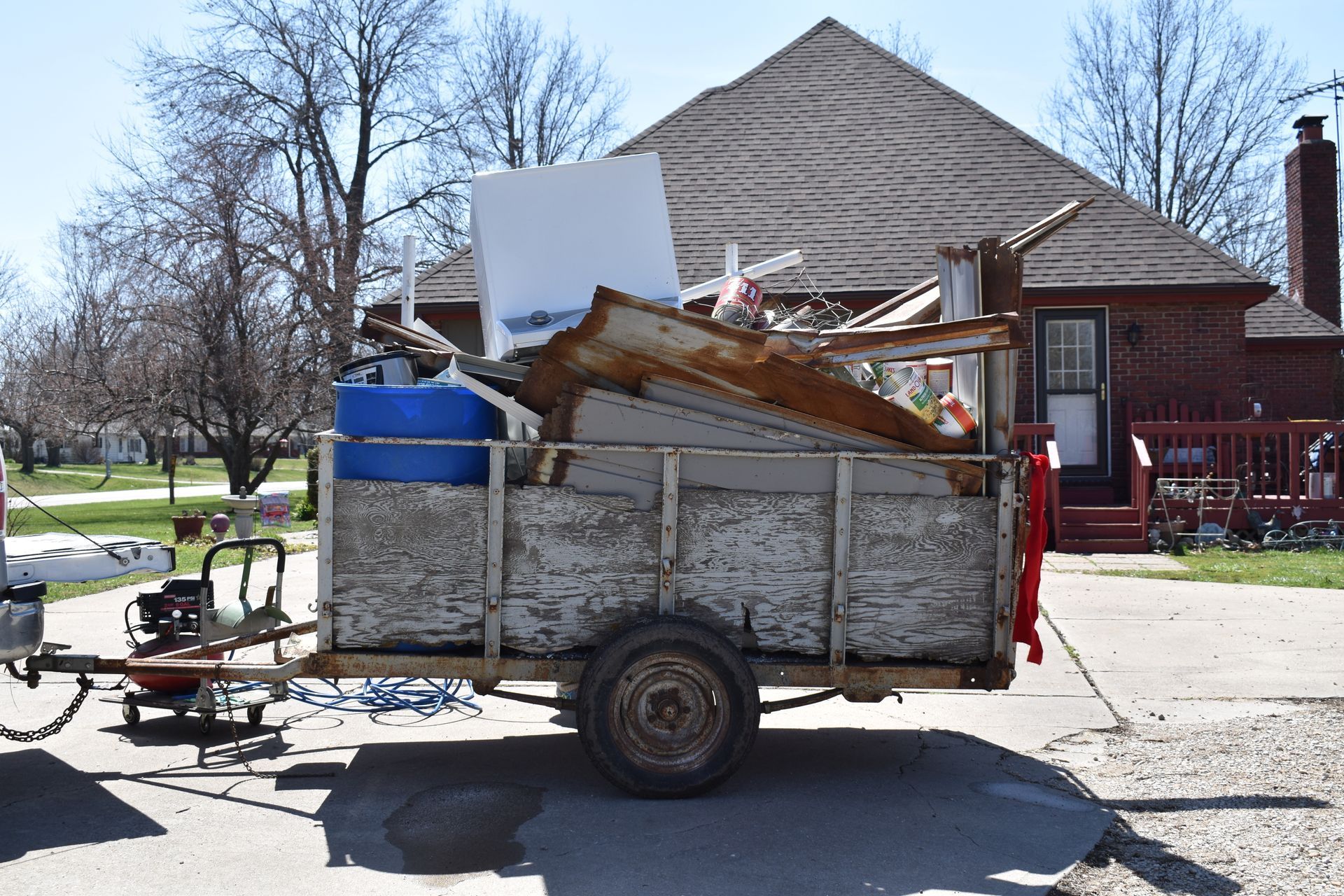 A utility trailer packed with debris and a blue plastic barrel, parked on a paved driveway in front of a brick house.