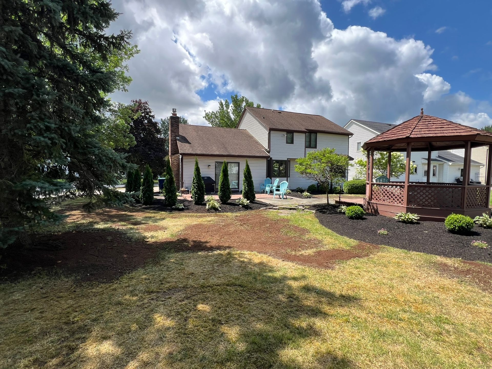A residential backyard featuring a two-story home, a wooden gazebo, manicured trees, and a lawn under a cloudy blue sky.