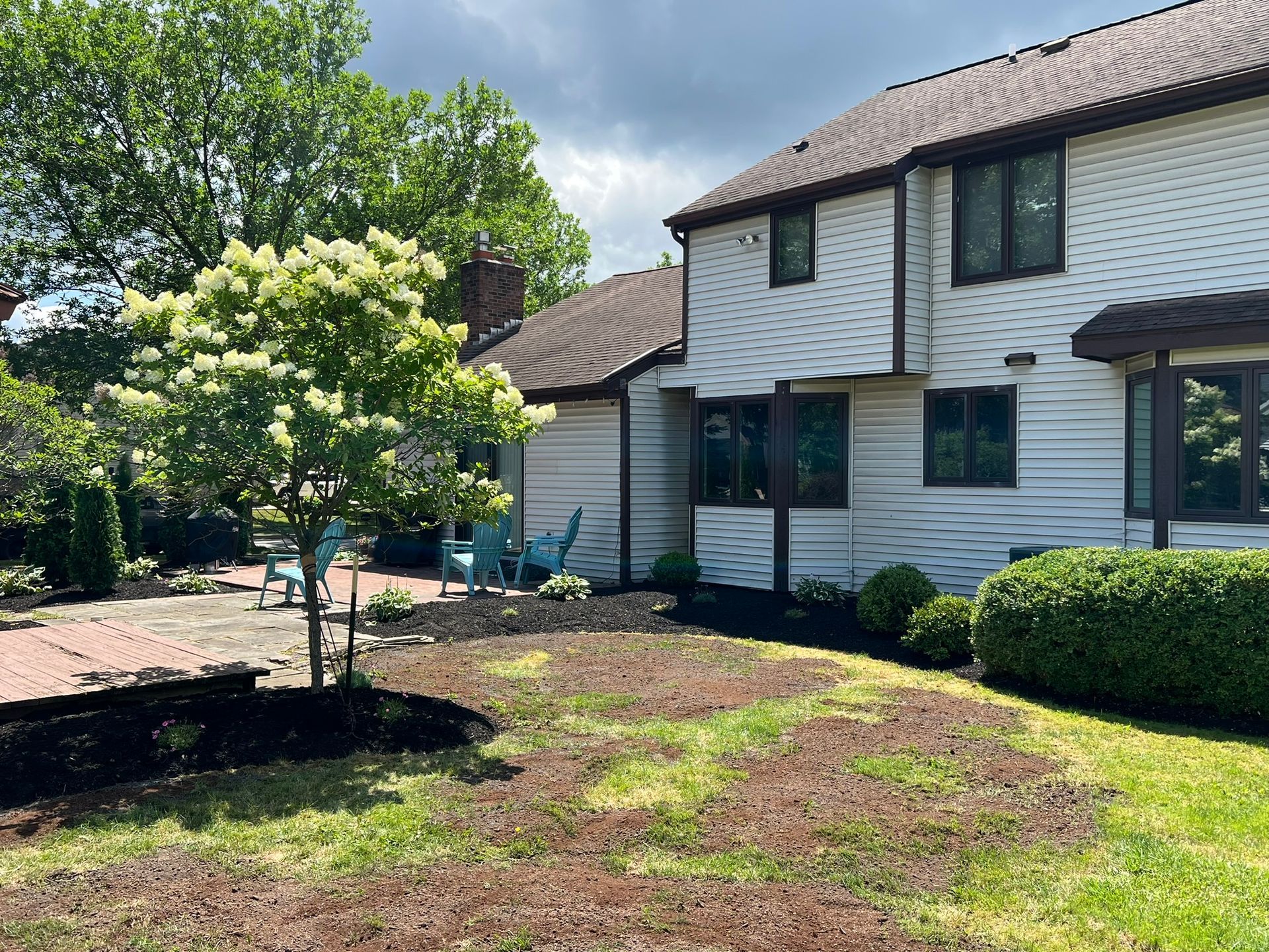 A white, multi-story house with a backyard patio featuring a flowering tree, garden beds, and blue outdoor chairs.