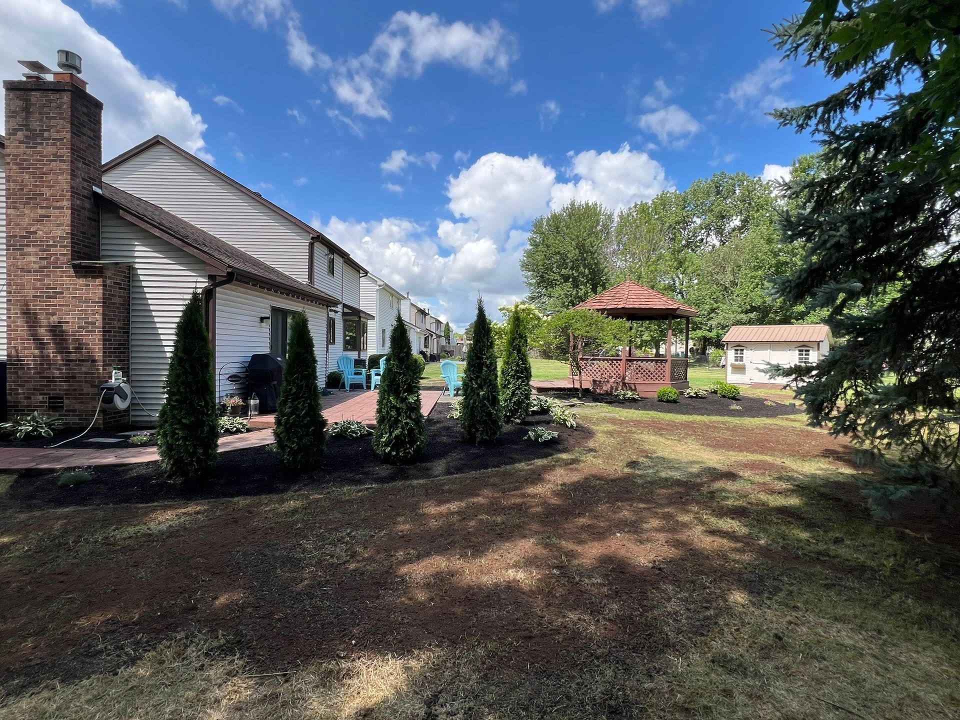 A backyard scene featuring a white house with a brick chimney, a brick patio, a wooden gazebo, and several small pines.