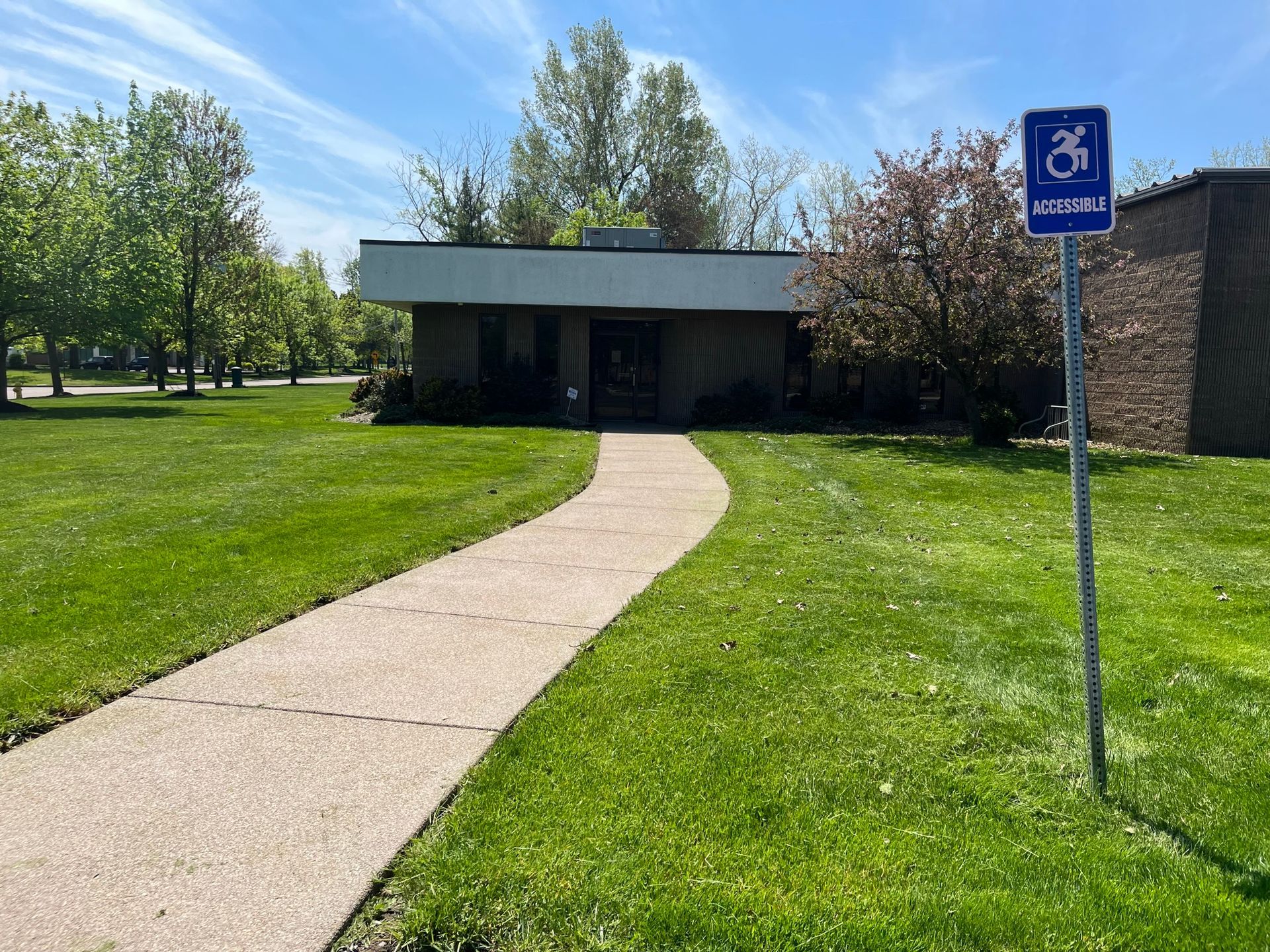 A sidewalk leads to the entrance of a flat-roofed building surrounded by green grass and trees, with a blue handicap sign.