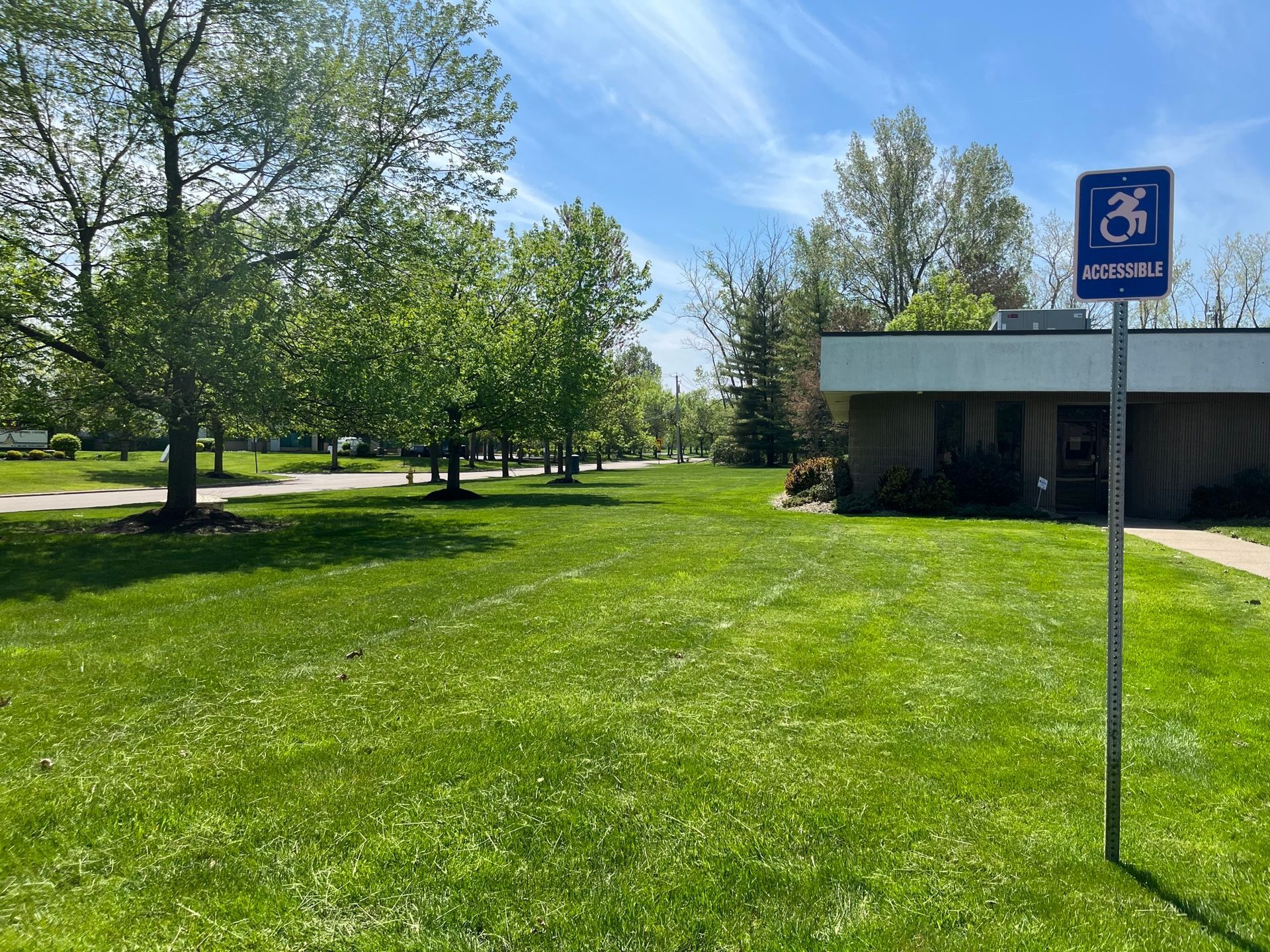 A blue handicap parking sign stands on a metal pole in a grassy lawn next to a building under a bright blue sky.