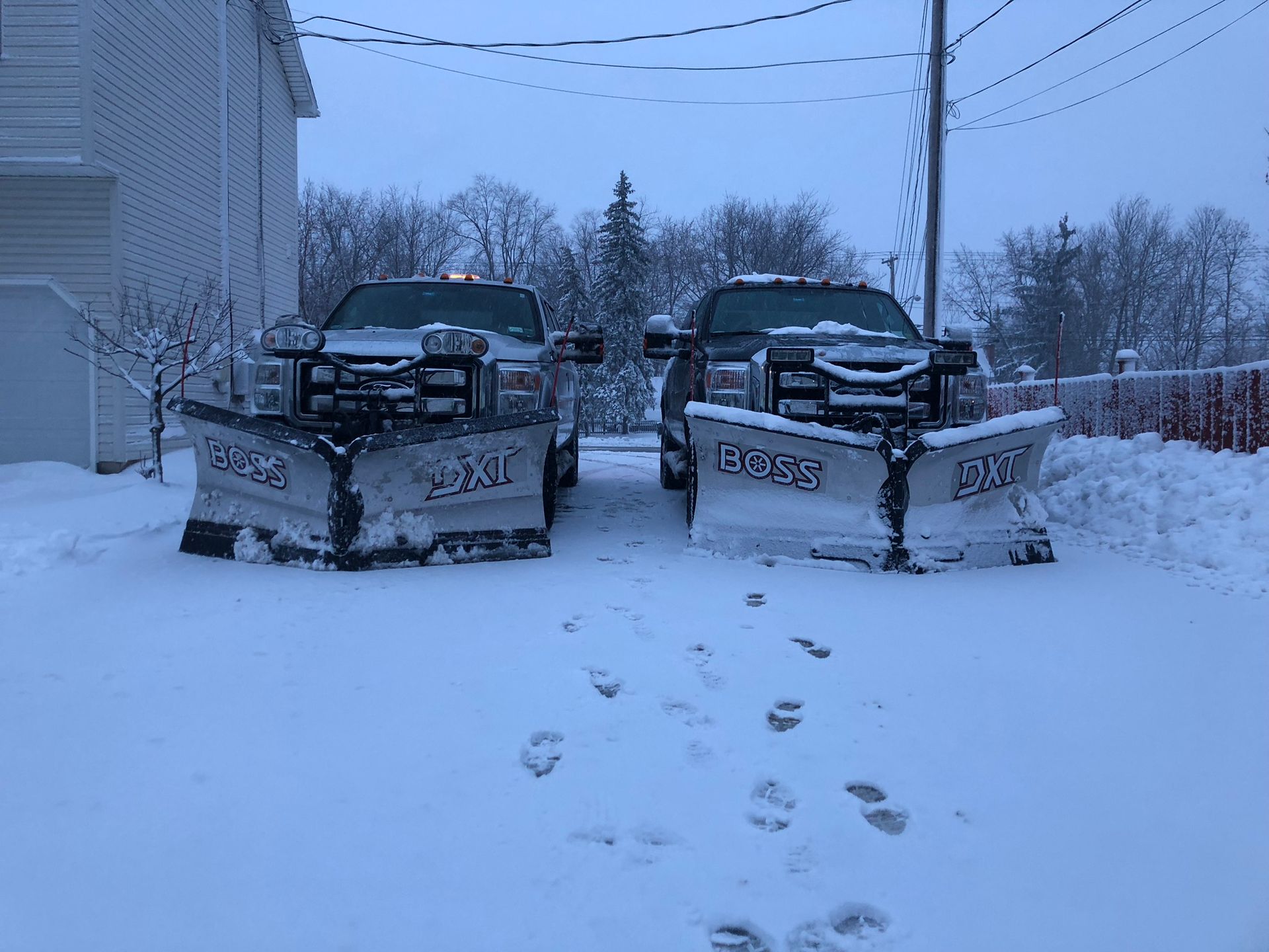 Two pickup trucks equipped with snowplows parked side-by-side in a snow-covered driveway during a winter storm.