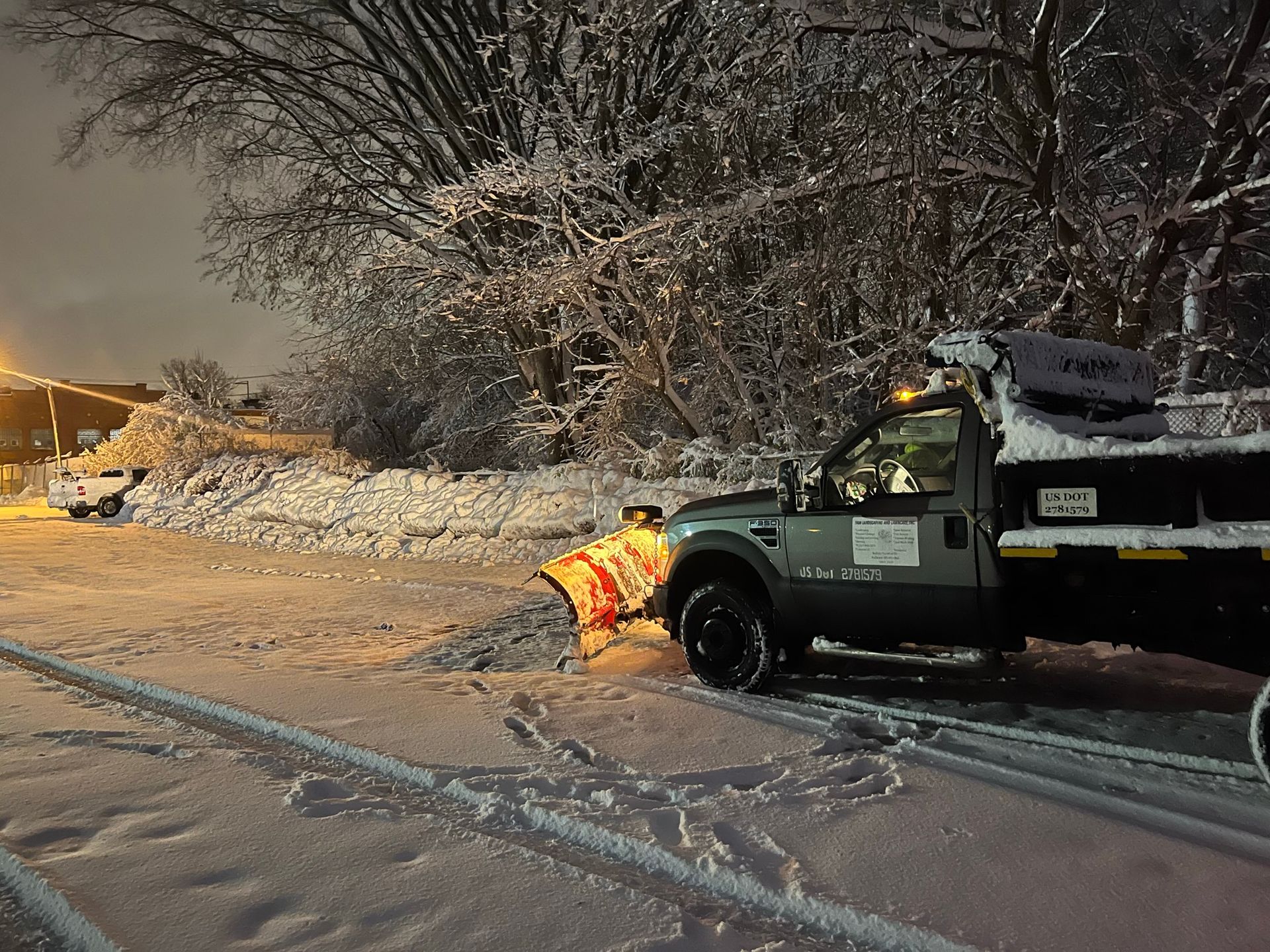 A work truck with a snow plow clears snow from a road at night under snow-covered trees and a streetlamp.