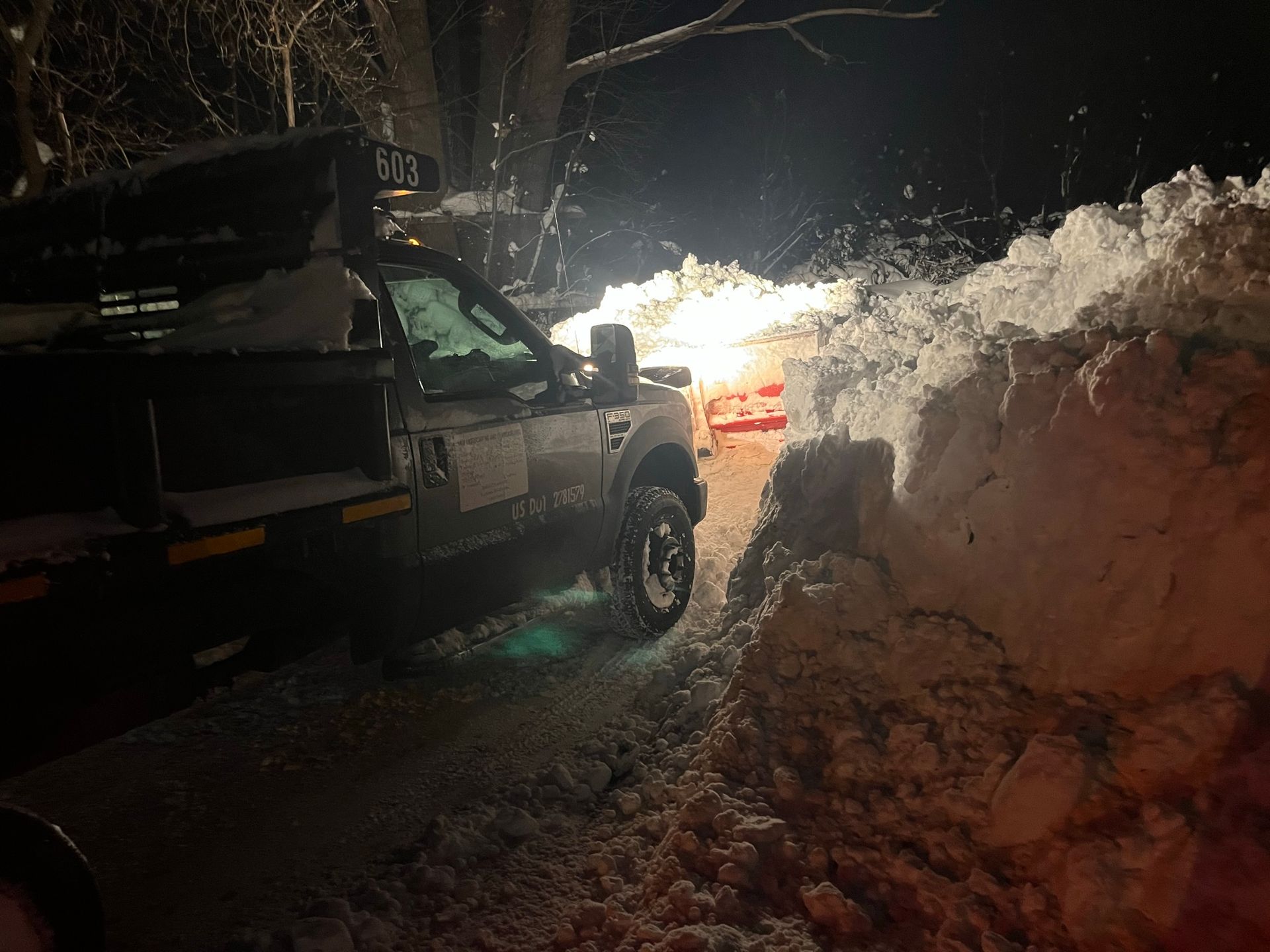 A snowplow truck travels along a road at night, surrounded by tall banks of snow.