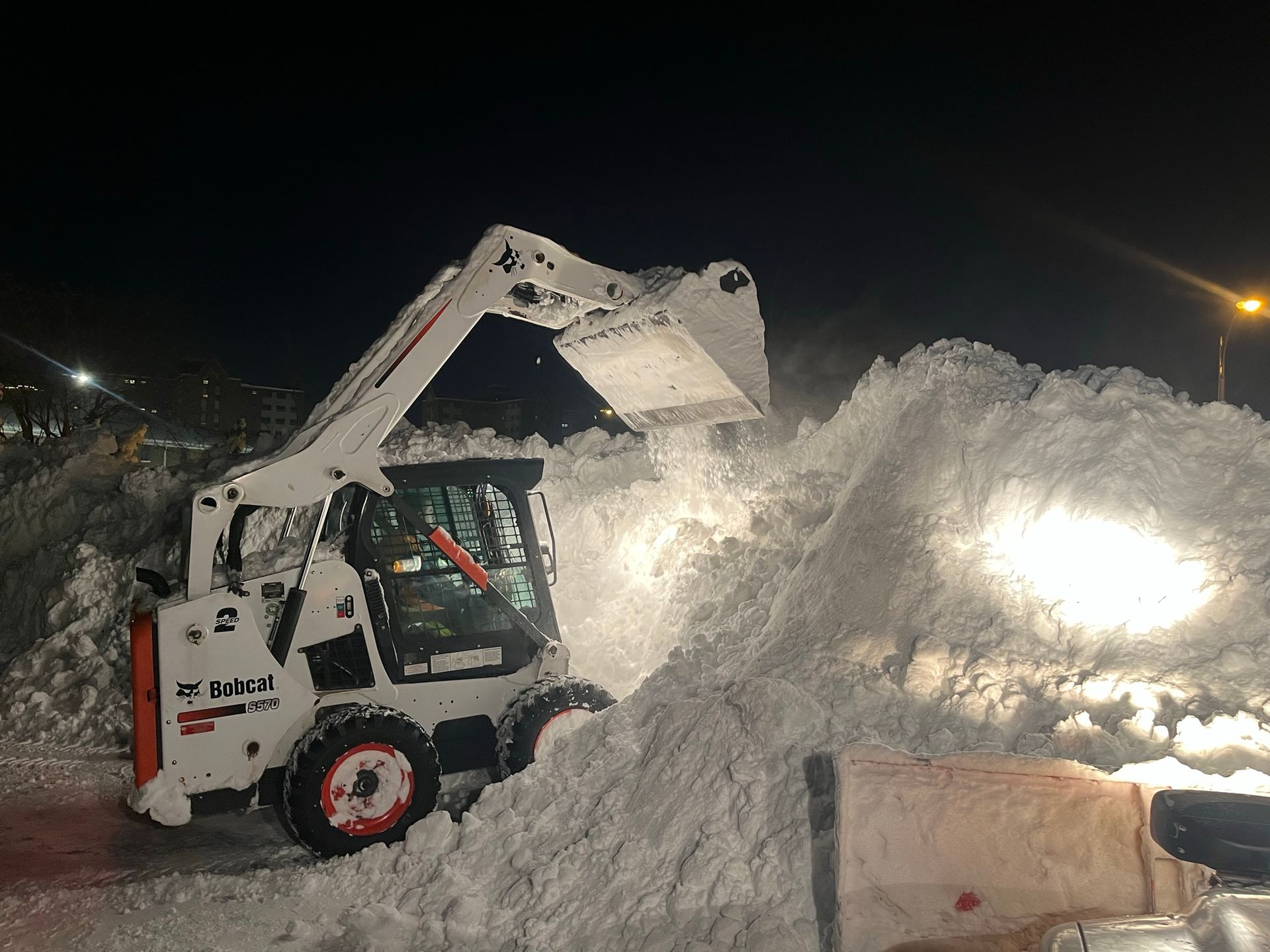 A white Bobcat skid-steer loader clears a large pile of snow at night under bright work lights.