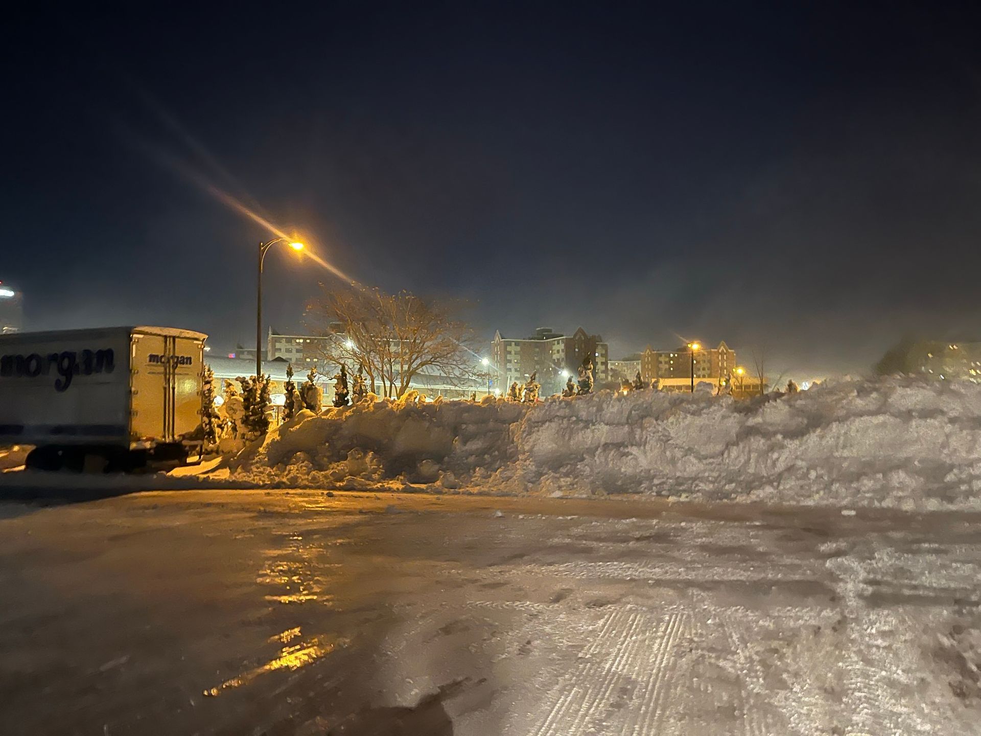 A night scene of a parking lot covered in ice with large snow piles, a parked box truck, and bright street lighting.