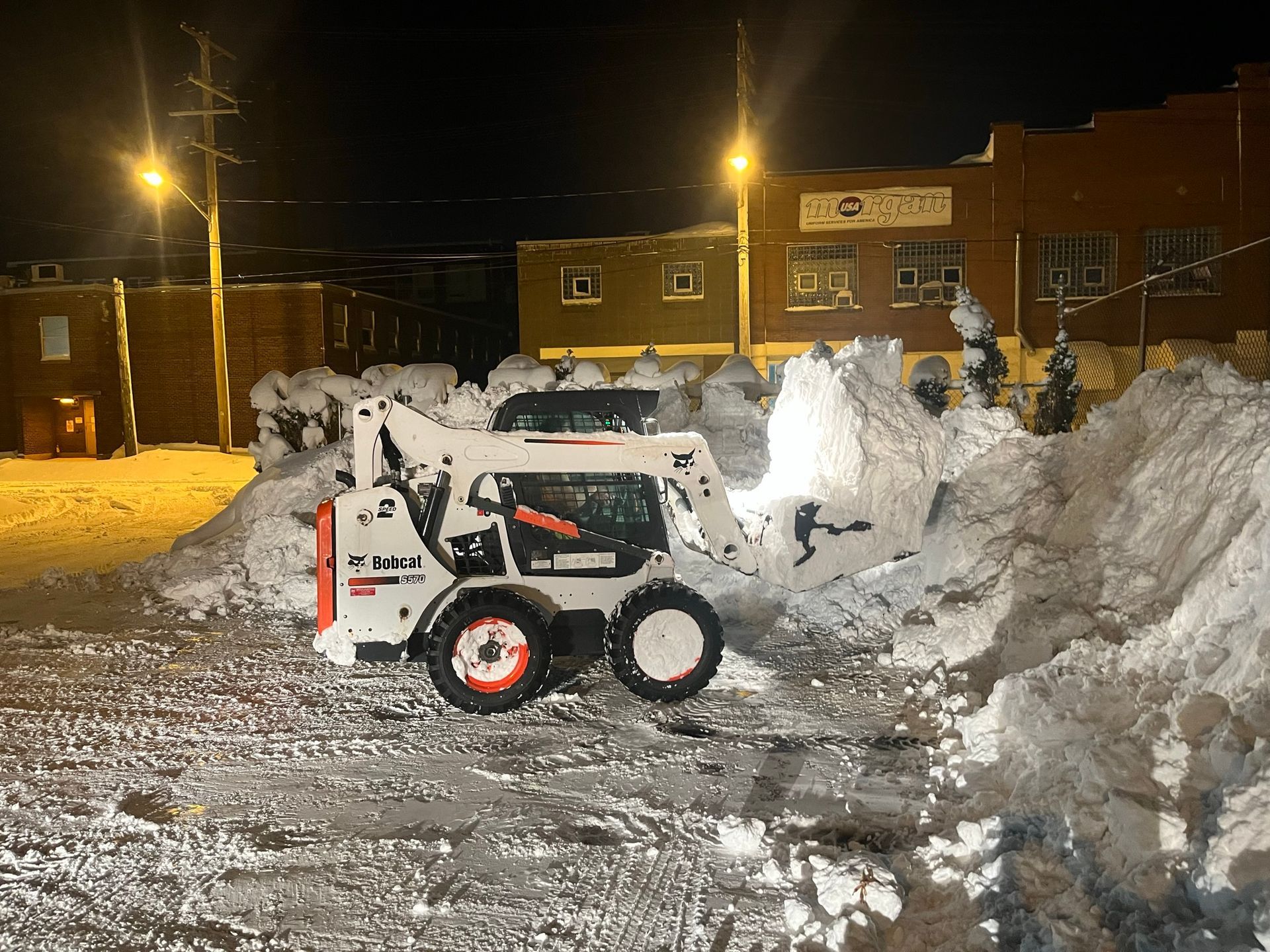 A white Bobcat skid-steer loader piles snow in a parking lot at night under bright streetlights.