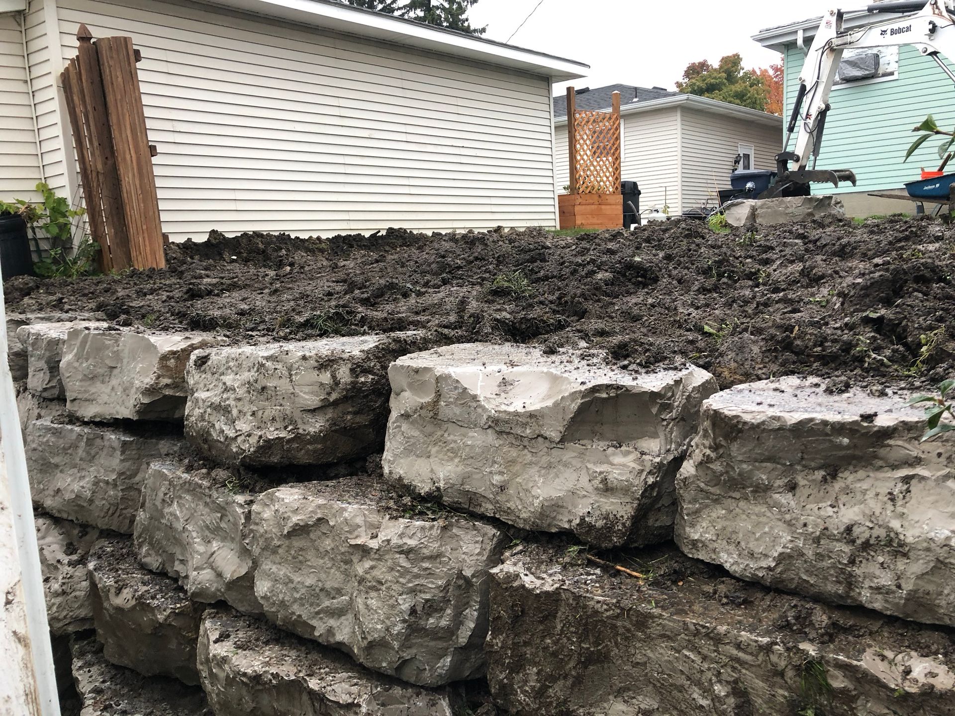 A stacked stone retaining wall filled with dark soil in a backyard, with a residential house and construction equipment.