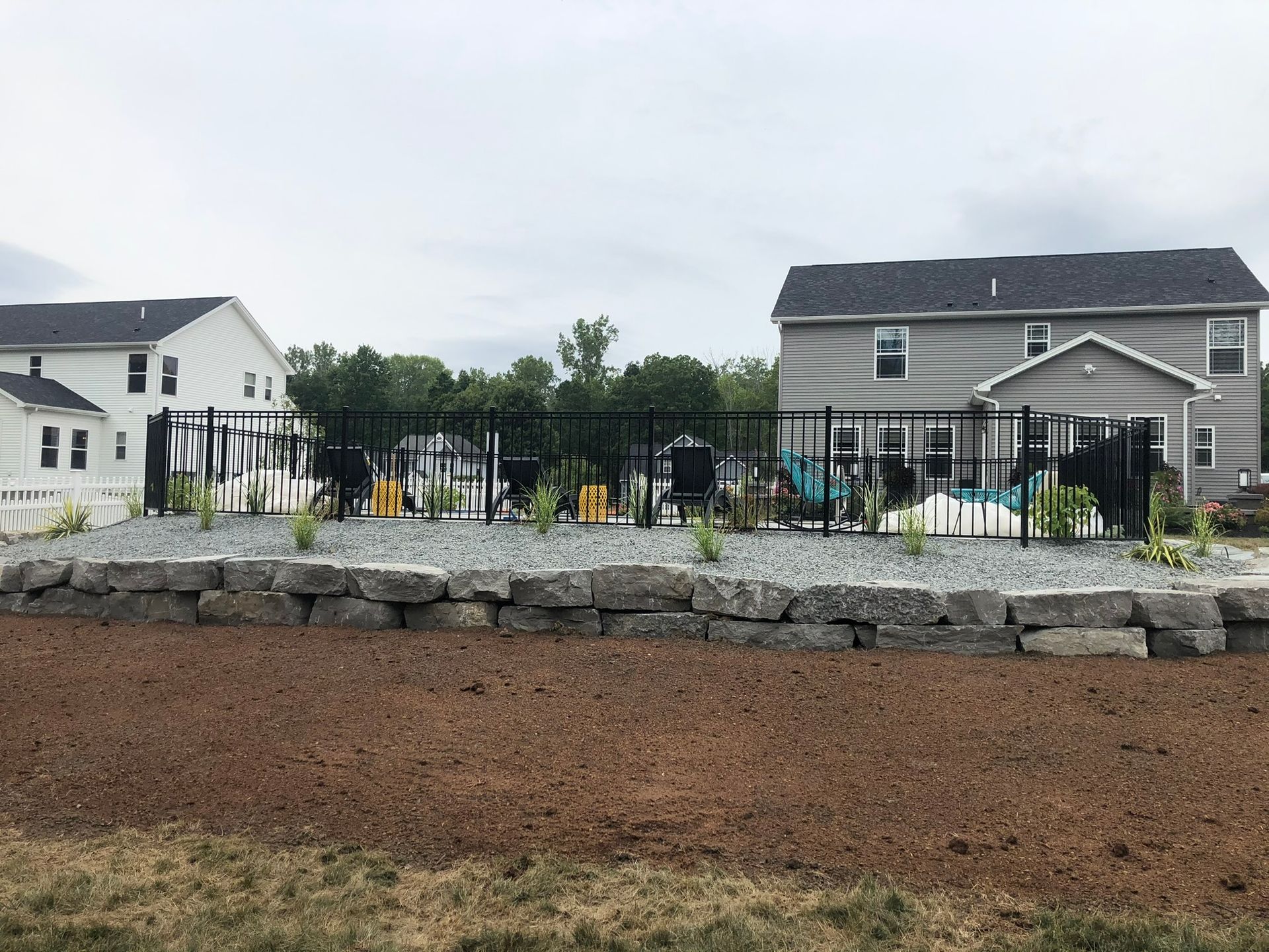 A tiered stone retaining wall separates a mulched yard from a fenced-in pool area between two suburban houses.