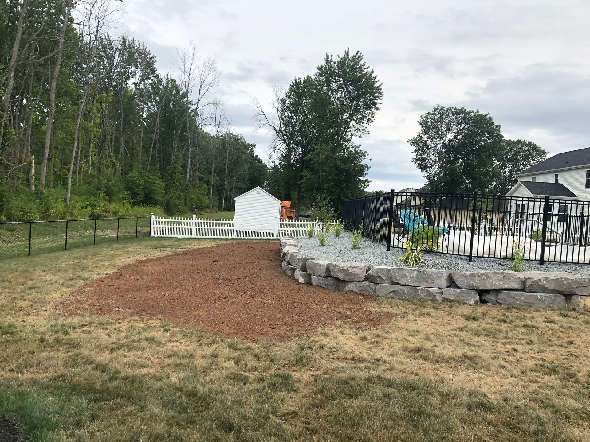 A backyard features a curved stone retaining wall, a patch of brown mulch, a white picket fence, and a distant forest.