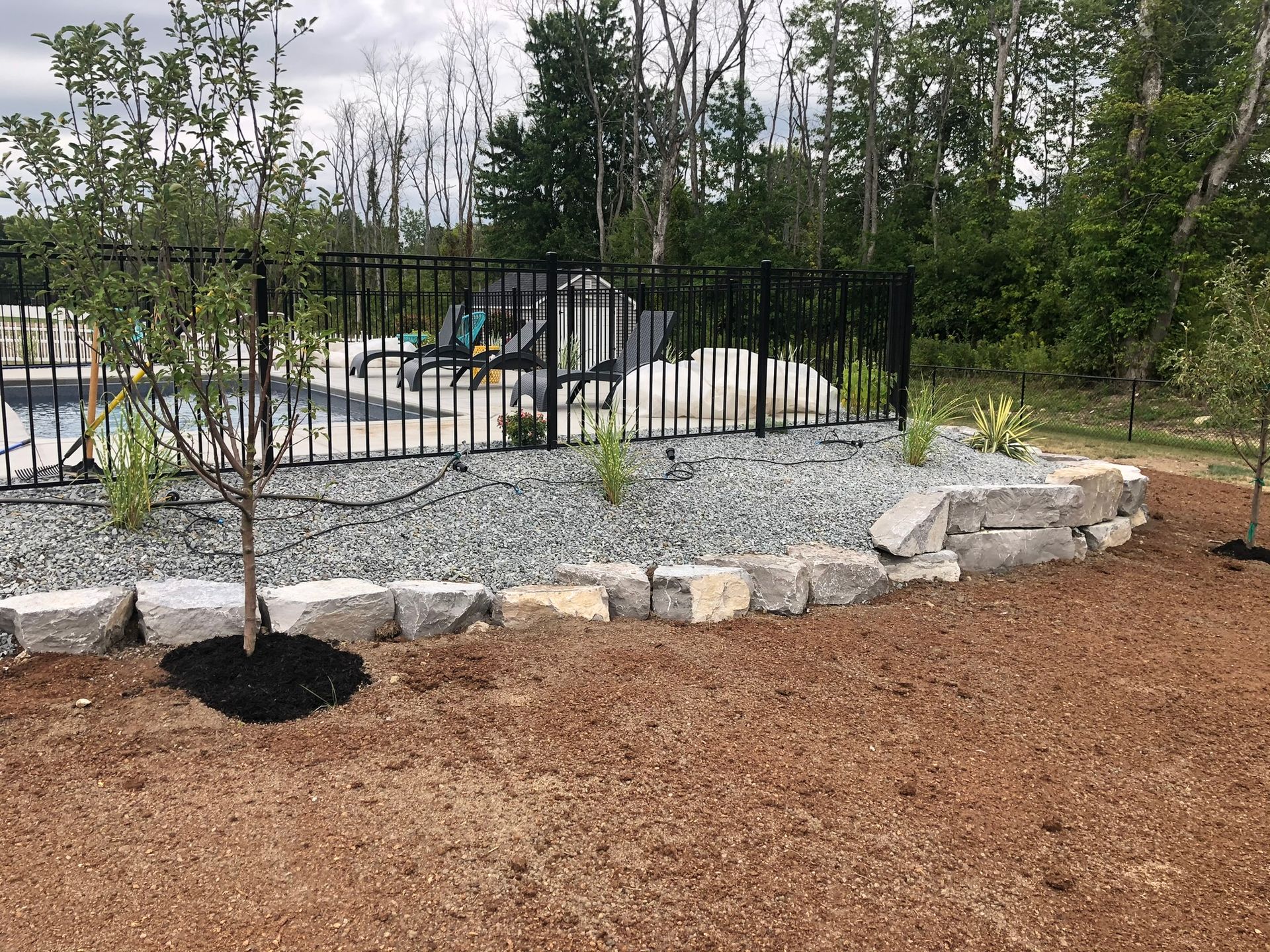 A stone retaining wall bordering a landscaped garden bed with a small tree in front of a fenced swimming pool area.