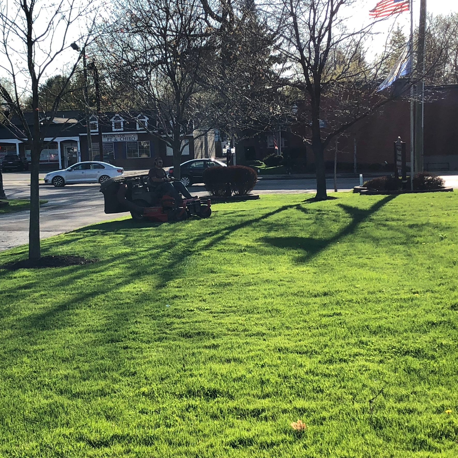 A person rides a lawnmower across a green park lawn on a sunny day with trees, a flag, and cars in the background.
