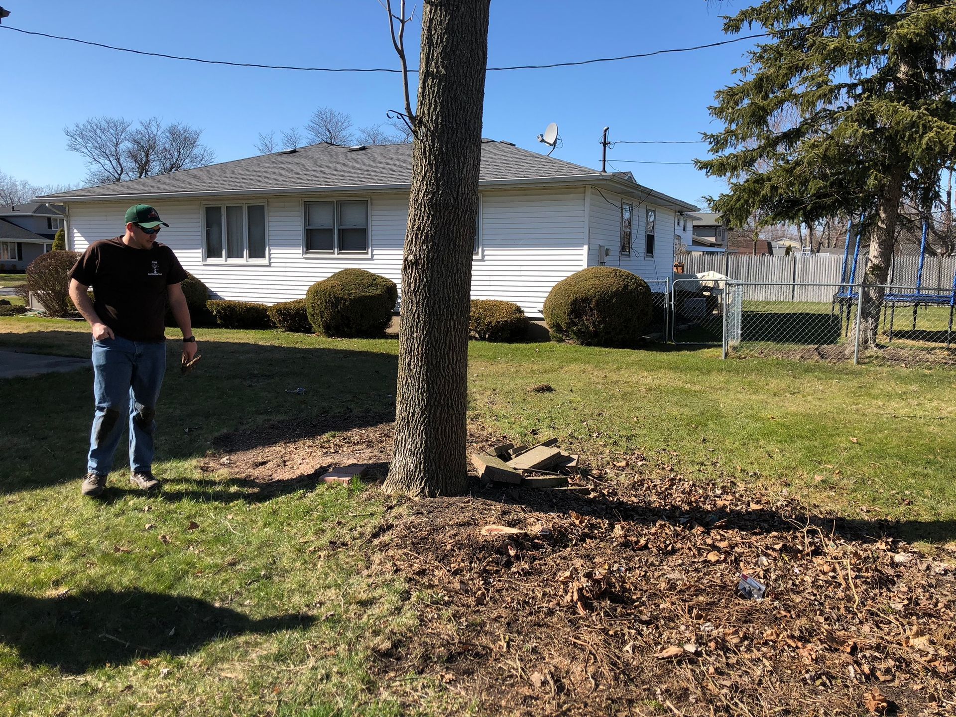 A person wearing a dark shirt and jeans stands on a grassy lawn next to a tree in front of a white house.