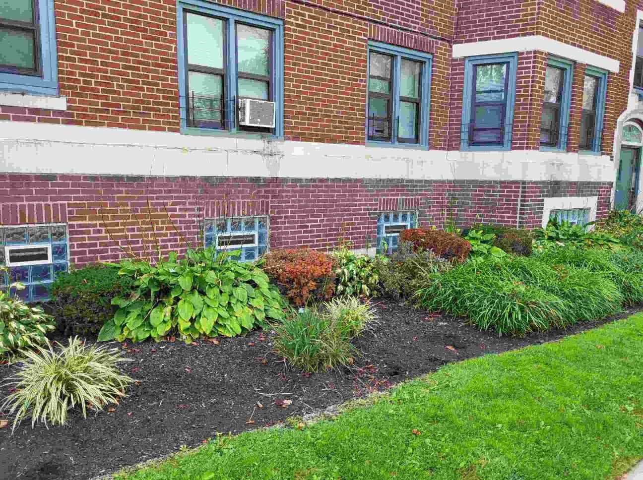 A brick apartment building exterior with foundation landscaping, glass block basement windows, and fresh mulch.