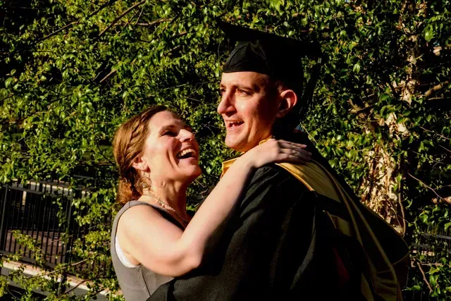 Smiling graduate in cap and gown embracing a woman outdoors by greenery