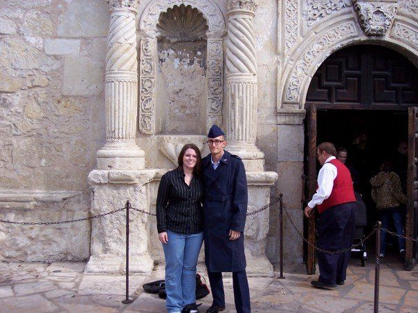 Two people posing in front of an ornate stone building entrance, with a man standing near the doorway.