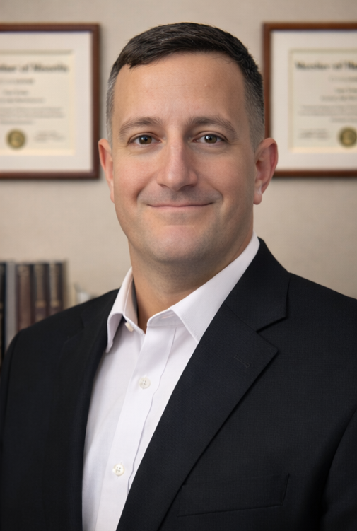 Man in black suit and white shirt, smiling, in front of diplomas.