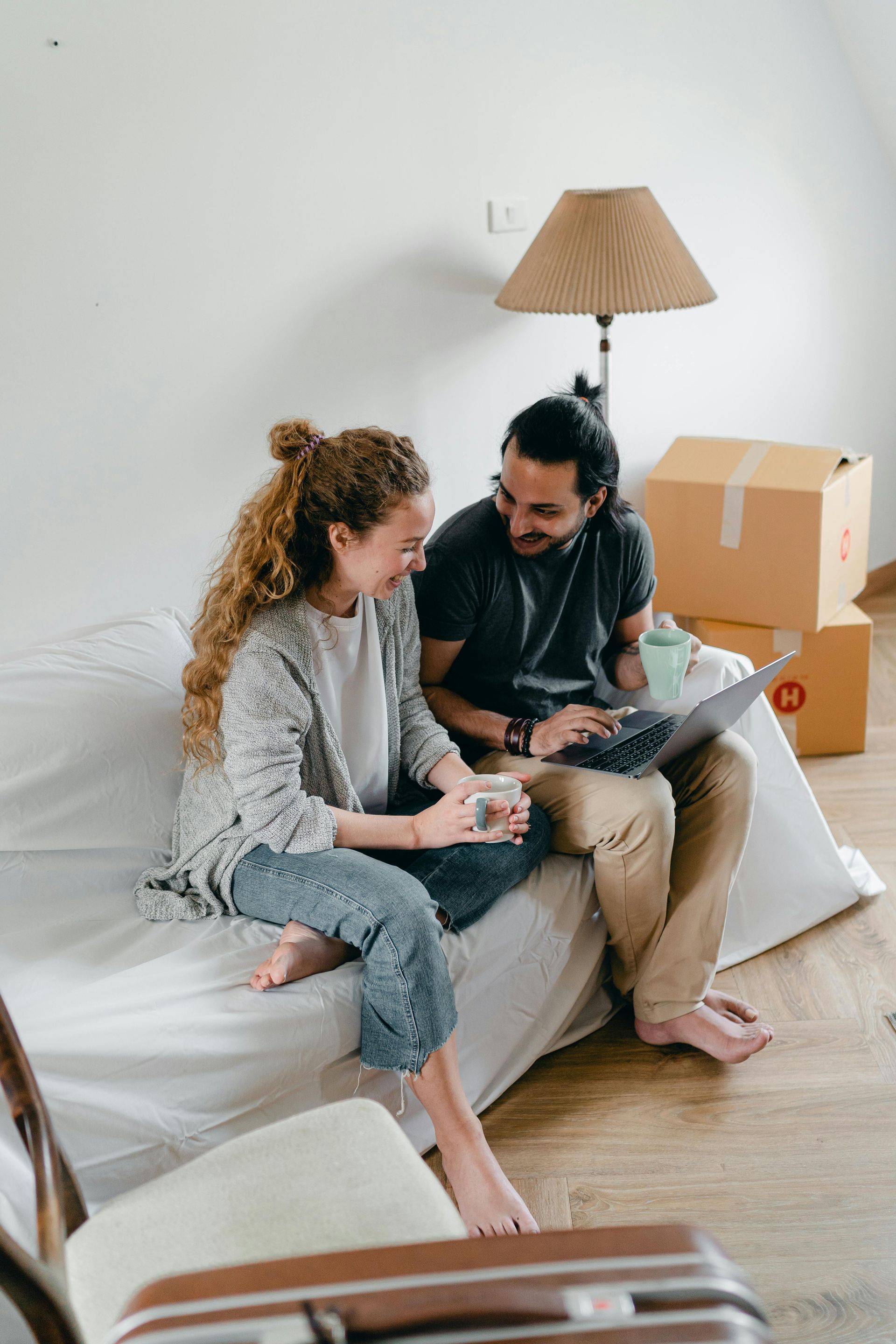 Couple sits on bed, smiling, looking at laptop. Moving boxes in background.