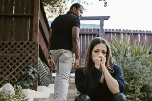 Man walking away from a woman sitting outside, covering her face with her hands, looking sad.