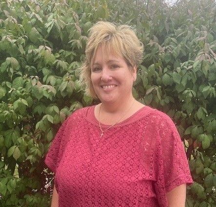 Woman with blonde hair wearing a red shirt, smiling, in front of leafy green bushes.