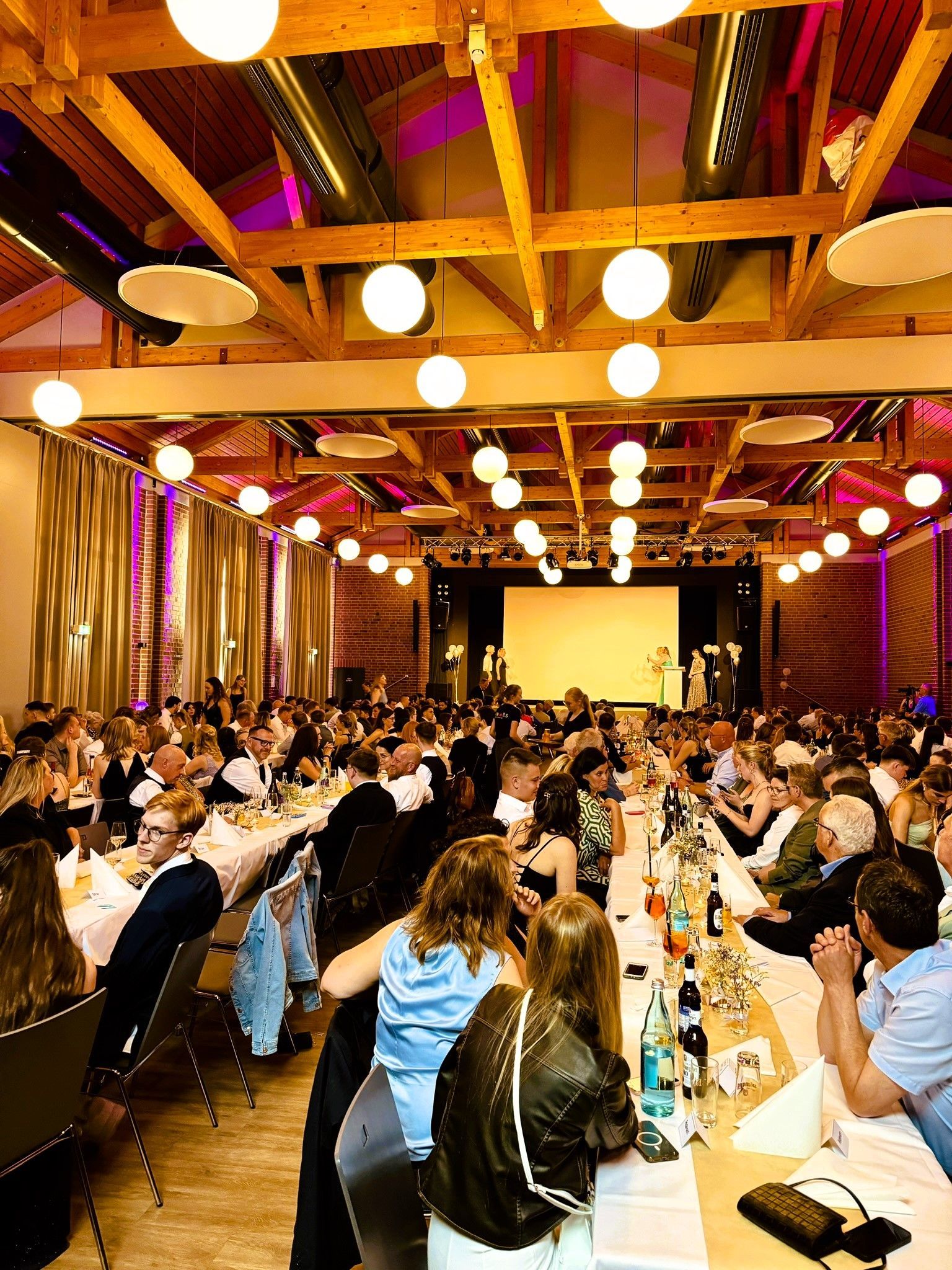Eine große Gruppe von Menschen sitzt an langen Tischen in einem großen Raum.