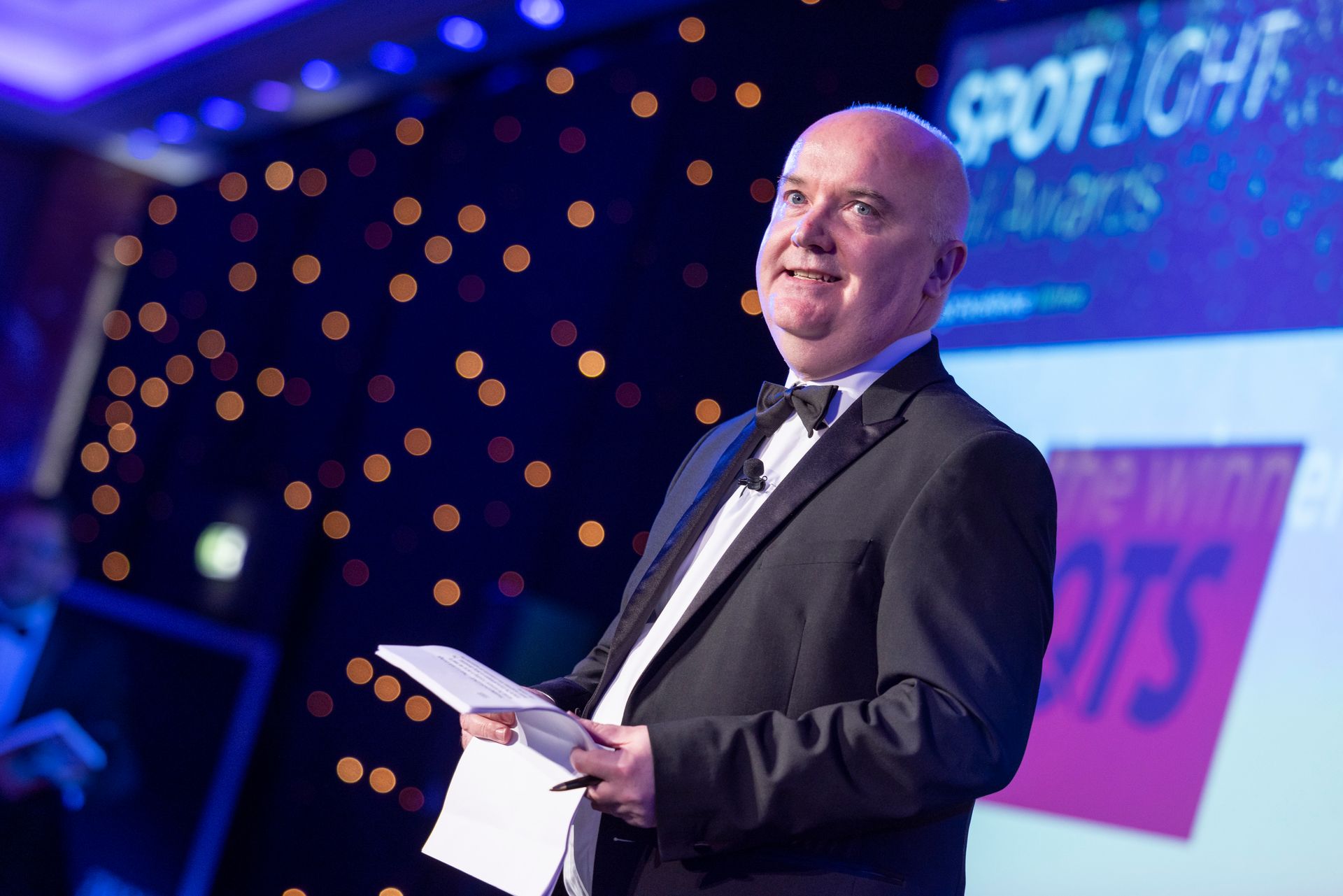 A man in a tuxedo is standing on a stage holding a piece of paper.
