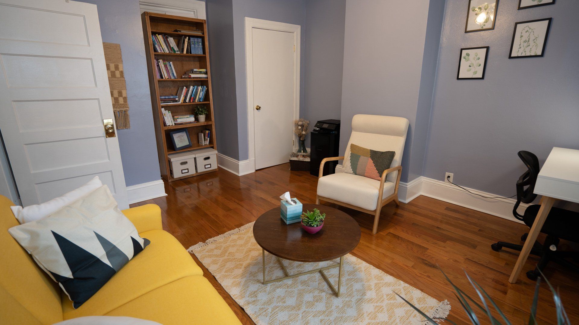 A therapy office  with a yellow couch , chair , coffee table and bookshelf.