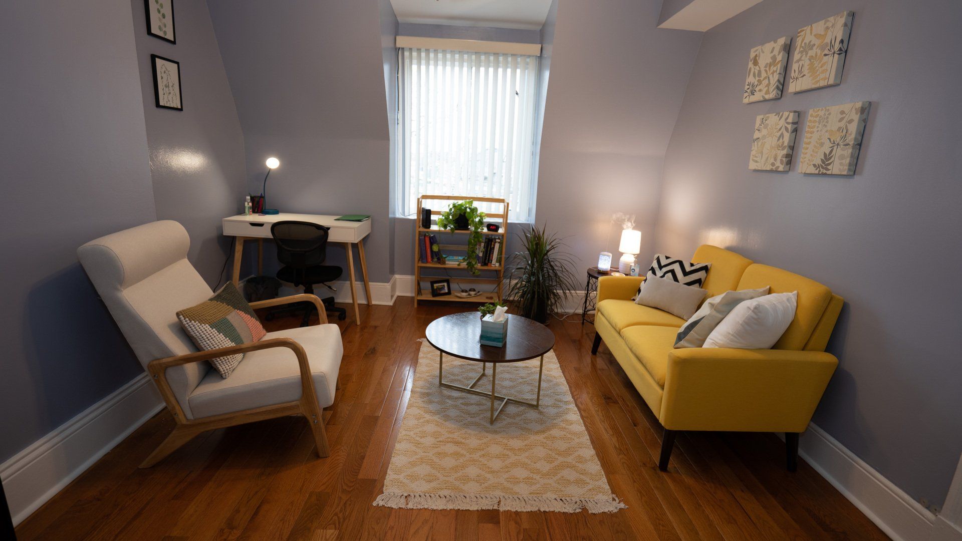 A therapy office with a yellow couch , chair , desk and coffee table.